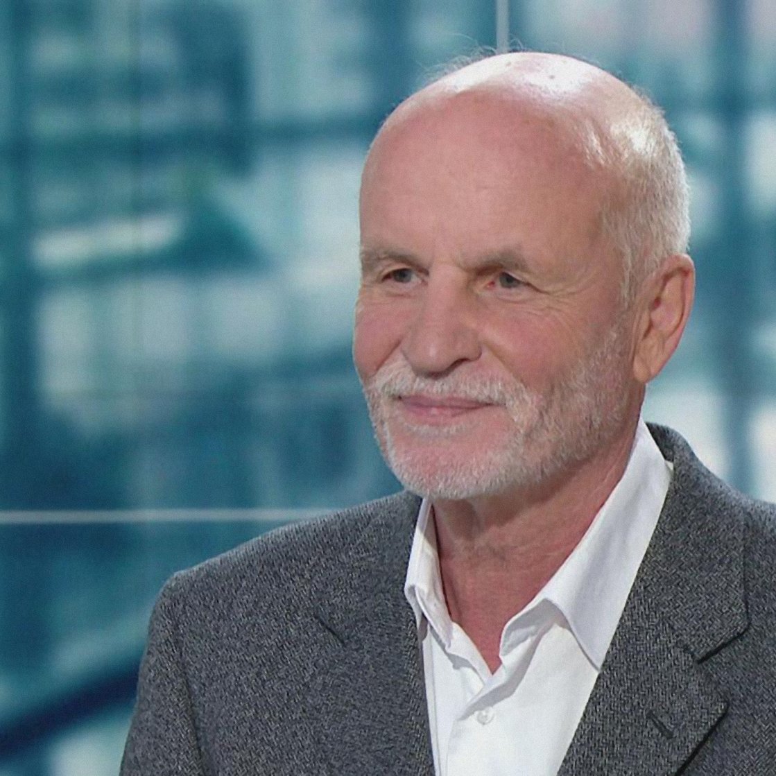 Homme âgé avec cheveux gris, souriant, portant un costume sobre sur fond flou.