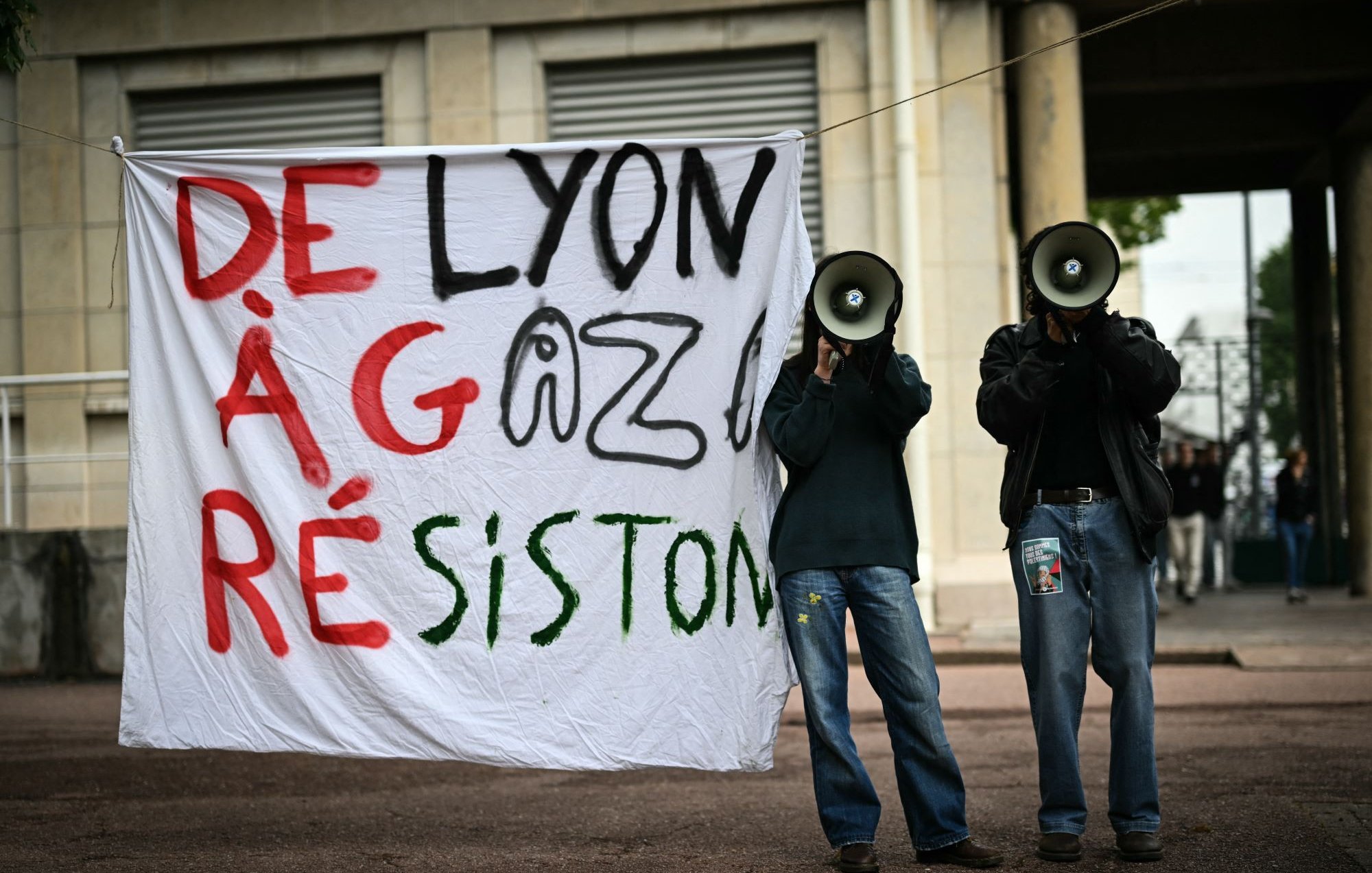 L'immagine mostra due persone che reggono un grande striscione bianco con le parole "DE LYON À GAZA RÉSISTON" scritte in caratteri grandi e colorati. Le due persone hanno in mano dei megafoni e sembrano partecipare a una manifestazione o a un evento di protesta. Lo sfondo suggerisce un contesto urbano, probabilmente in una zona pubblica.