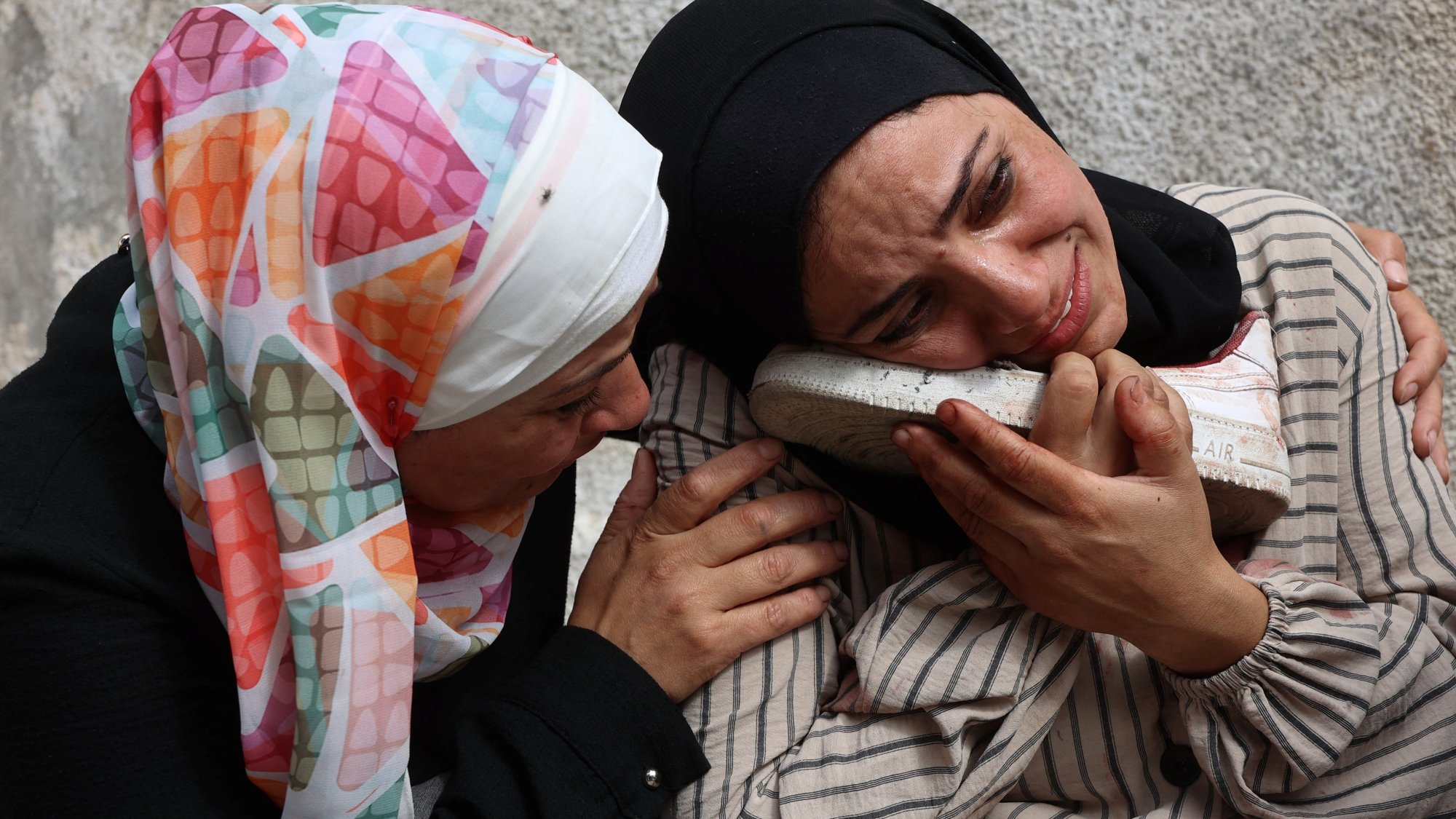 Due donne in abiti tradizionali si abbracciano, una piange e tiene un oggetto vicino al viso.
