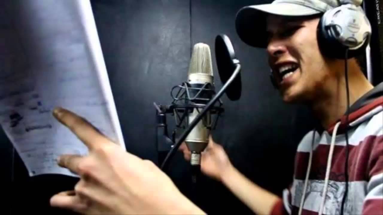 L'image montre un homme en train de chanter ou de rapper dans un studio d'enregistrement. Il porte un chapeau et des écouteurs. Devant lui, un microphone est connecté et il tient des feuilles de papier, probablement des paroles ou un texte. L'expression de son visage indique qu'il est très impliqué dans sa performance. L'arrière-plan semble sombre, ce qui est typique des environnements d'enregistrement.