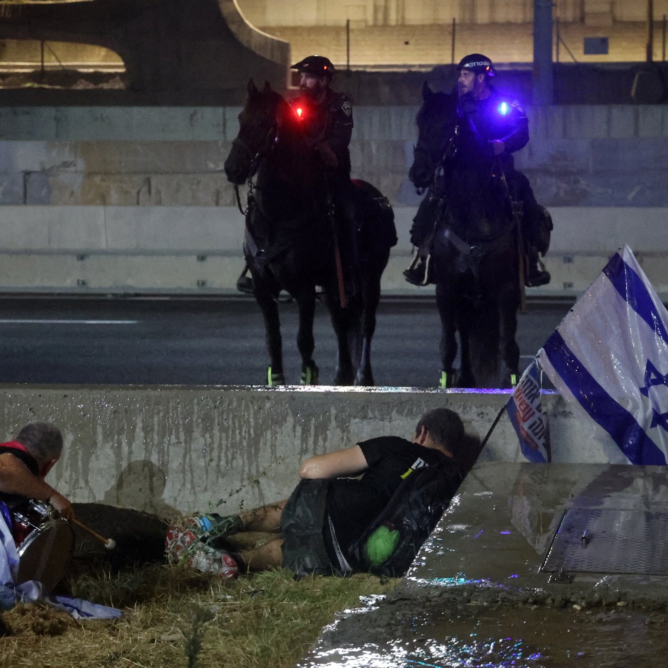 تظهر الصورة مشهدًا في مدينة ما، حيث يتواجد رجلان على الأرض بجانب ممر مائي أو خندق. أحدهما يبدو أنه على الأرض بينما الآخر يختبئ خلف حواجز. في الخلفية، يظهر اثنان من رجال الشرطة على ظهور الخيل، مجهزين بأضواء ملونة على خوذتهم. هناك أيضًا علم إسرائيلي ملقى على الأرض بالقرب من الرجلين. الجو العام يوحي بوجود حدث أو مواجهة تتعلق بالتوترات الاجتماعية أو السياسية.