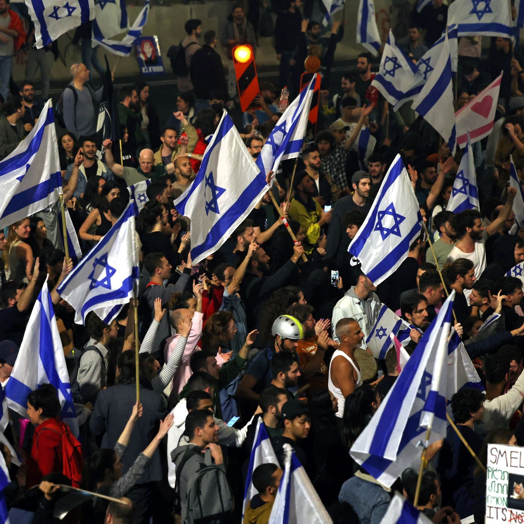 L'image montre une grande foule rassemblée, où des personnes tiennent des drapeaux israéliens. L'atmosphère est festive et engagée, avec des signes et des expressions de mobilisation. On peut observer une diversité de participants, certains portant des cris ou des slogans. La scène semble se dérouler lors d'une manifestation ou d'un rassemblement public.