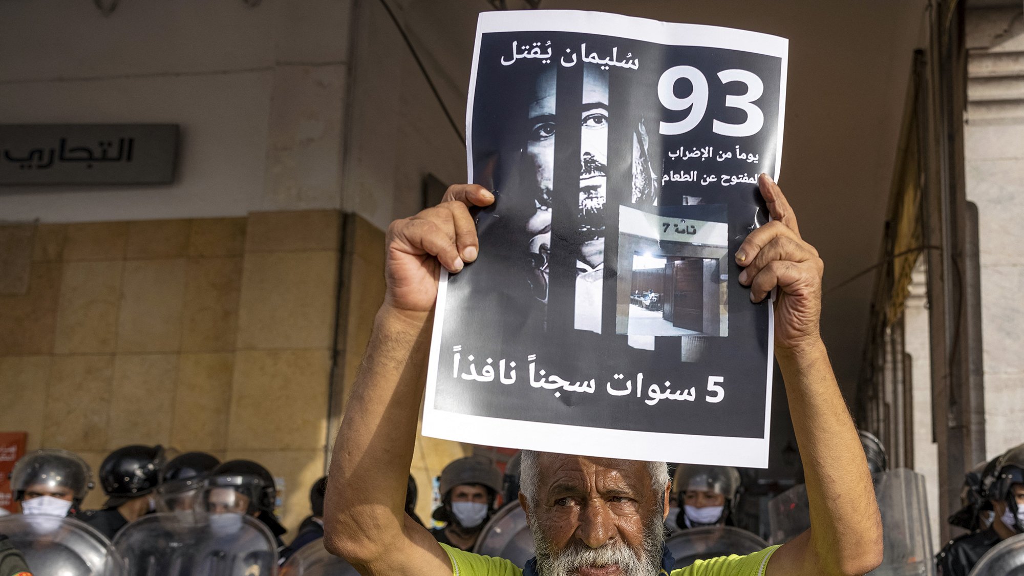 L'image montre un homme âgé tenant une pancarte en arabe. La pancarte mentionne des chiffres et des phrases qui semblent évoquer une question de justice ou de prison. En arrière-plan, on aperçoit des policiers en tenue d'émeute, ce qui suggère une atmosphère de protestation ou de tension. L'homme exprime probablement un sentiment d'injustice ou de revendication sociale.