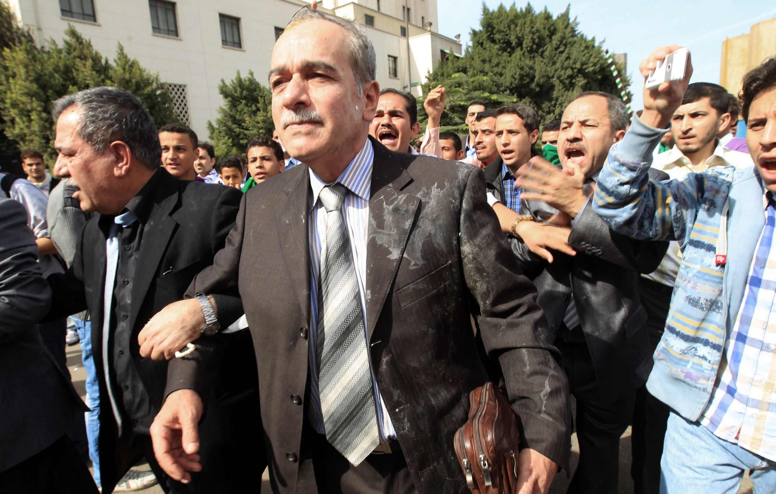 L'image montre un groupe de personnes dans une scène dynamique et animée. Au centre, un homme en costume, portant une cravate, semble marcher avec détermination, bien qu'il ait des éclaboussures sur son vêtement. Il est entouré de plusieurs autres individus qui semblent agités et expriment des émotions fortes, peut-être en criant ou en gesticulant. L'arrière-plan montre un environnement urbain avec des bâtiments et des arbres. L'atmosphère semble chargée, suggérant une situation de tension ou de protestation.
