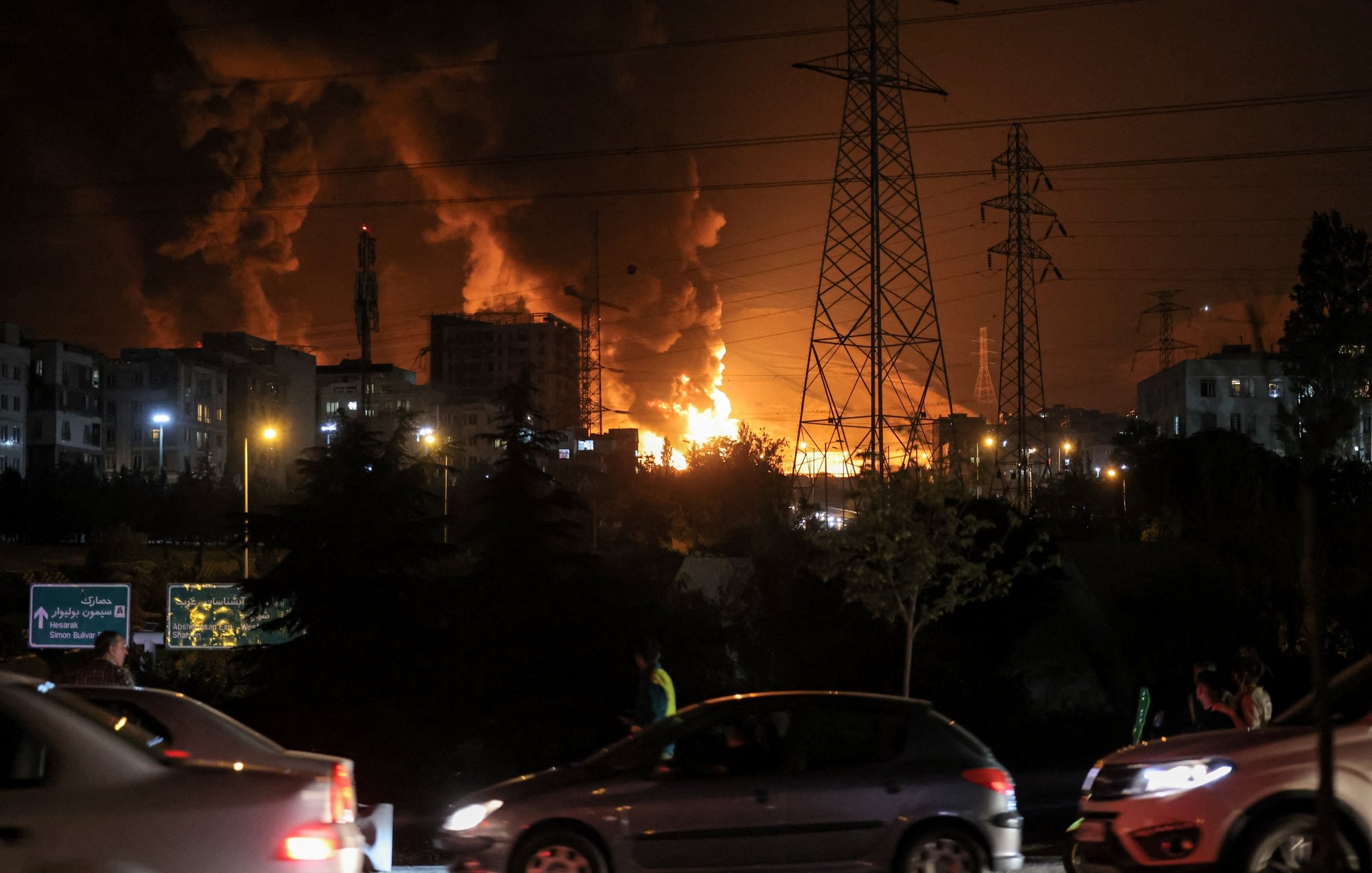 Bright flames and dark smoke rise from buildings at night, with cars passing nearby.
