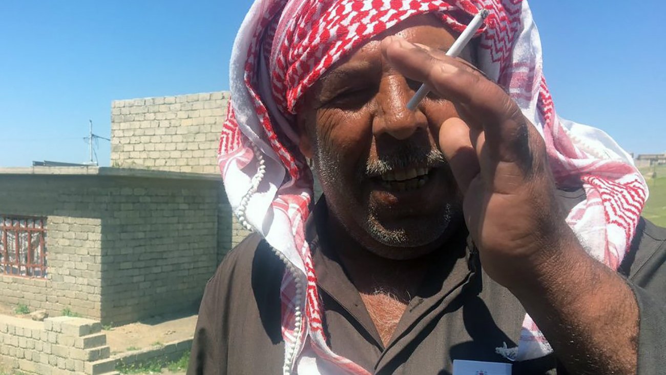 The image shows a man wearing a traditional headscarf, often referred to as a keffiyeh or ghutrah, with a red and white pattern. He is standing outdoors, possibly in a rural area, with stone structures visible in the background. The man appears to be smiling or in a candid moment, holding a cigarette between his fingers. The setting is bright, indicating a sunny day, and the landscape around him includes some greenery and dirt paths.