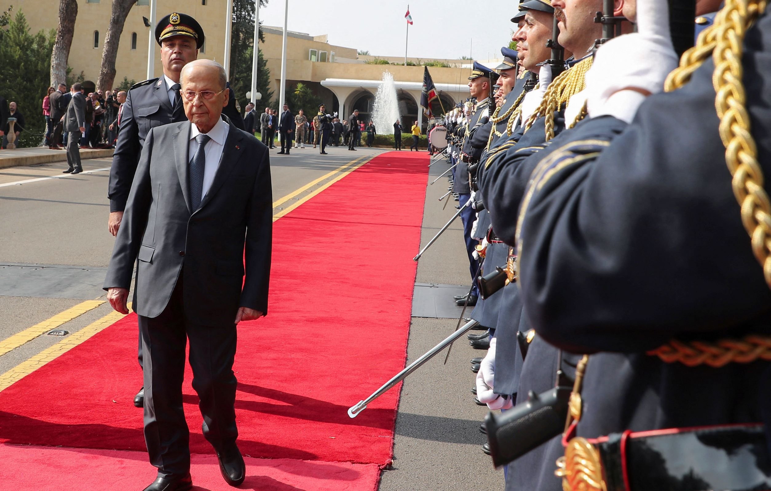 در این تصویر، یک مرد مسن با کت و شلوار تیره در حال راه رفتن بر روی فرش قرمز است. دو طرف او، گروهی از سربازان در لباس‌های رسمی و با تفنگ‌های خود ایستاده‌اند و به او احترام می‌گذارند. در پس‌زمینه، یک ساختمان رسمی و عناصر معماری نمایان است. به نظر می‌رسد که این مراسم یک استقبال رسمی باشد.