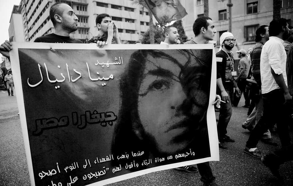 L'image montre une manifestation où des personnes portent une grande banderole. Sur la banderole, on peut voir le portrait d'une personne accompagnée d'un texte écrit en arabe, qui semble rendre hommage à cette personne. L'ambiance est chargée d'émotion, avec des manifestants en arrière-plan, et l'image est en noir et blanc, ce qui accentue la gravité de la situation. Les expressions des participants suggèrent une forte implication dans la cause défendue.