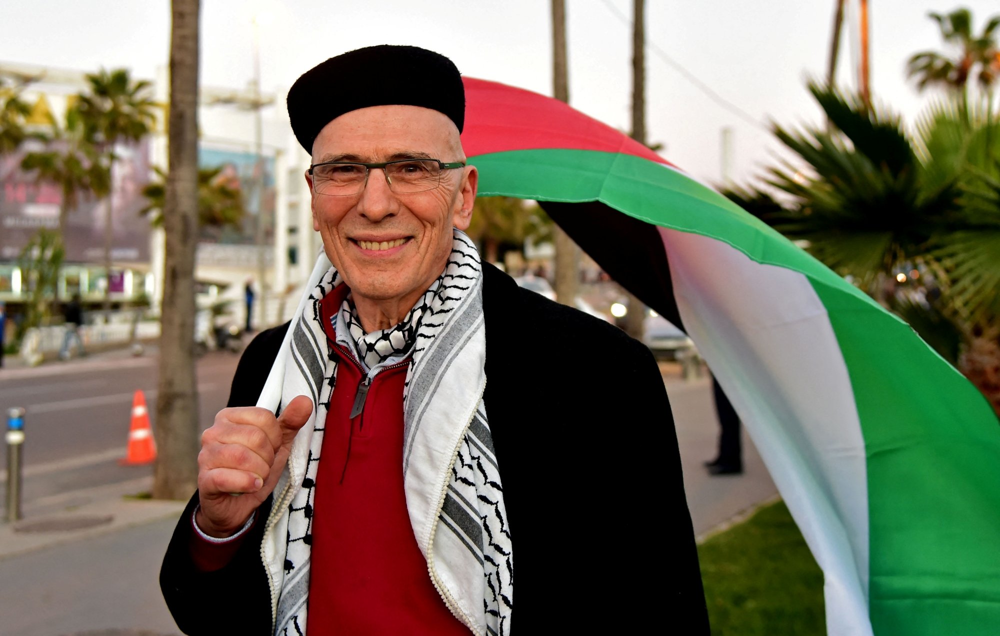 Un homme souriant porte un keffieh, derrière lui un drapeau palestinien sur fond urbain.
