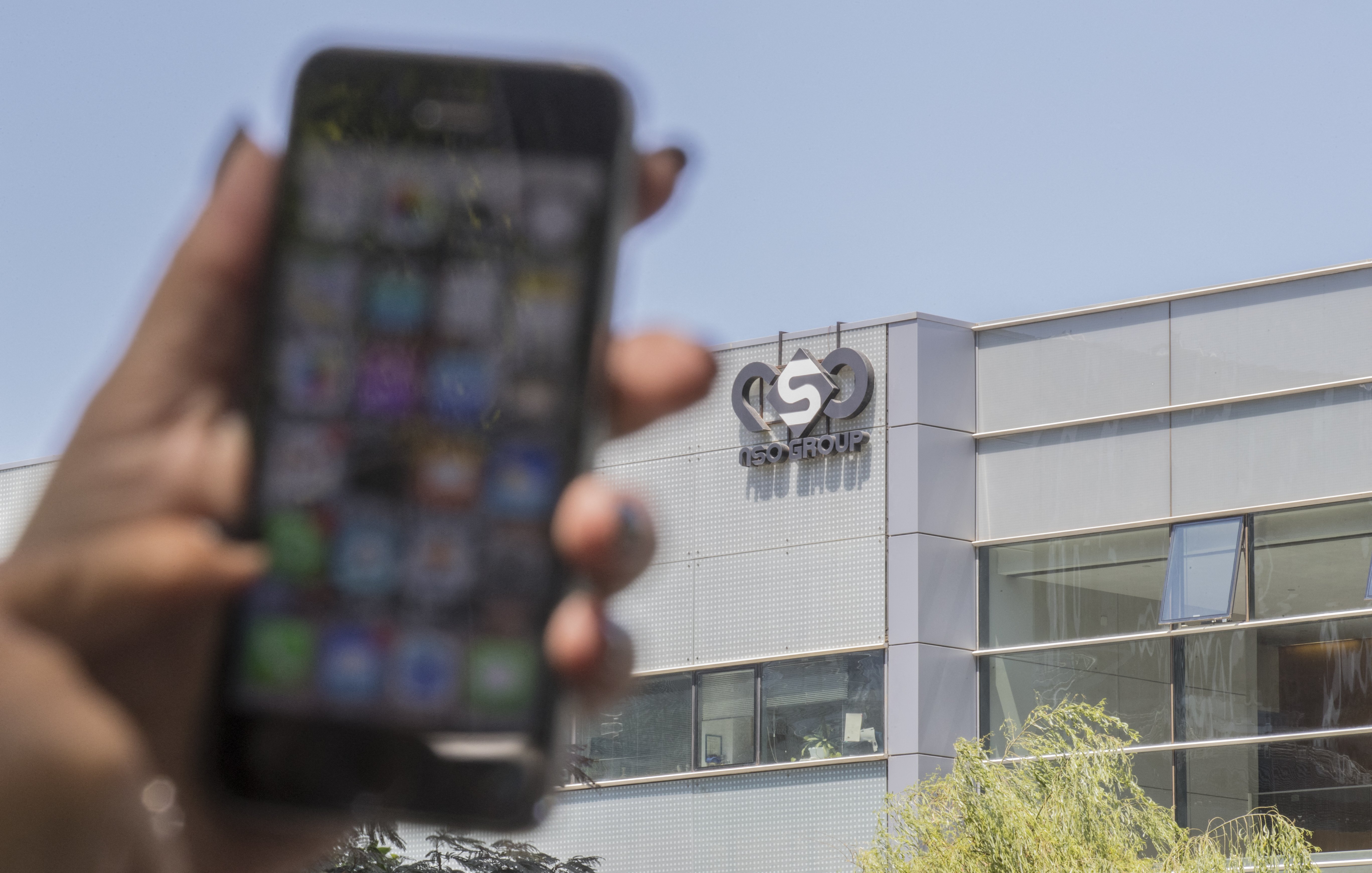 L'image montre un smartphone tenu par une main, flou au premier plan, avec un écran affichant diverses applications. En arrière-plan, on peut voir le bâtiment d'une entreprise, identifié par un logo qui semble être celui d'un groupe nommé "ISN GROUP". Le bâtiment a une façade moderne, probablement en verre. La lumière naturelle éclaire la scène, indiquant qu'il fait jour.