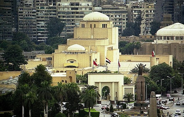 L'image montre un paysage urbain, probablement d'une grande ville. On peut voir des bâtiments aux architectures variées, certains avec des motifs modernes et d'autres avec des influences traditionnelles. Au centre, un bâtiment notable a un style distinctif avec un dôme et des éléments ornementaux, entouré de verdure. Des drapeaux flottent, ce qui suggère l'importance de cet endroit. En arrière-plan, des immeubles d'habitation se dressent, illustrant le contraste entre les différentes époques architecturales.