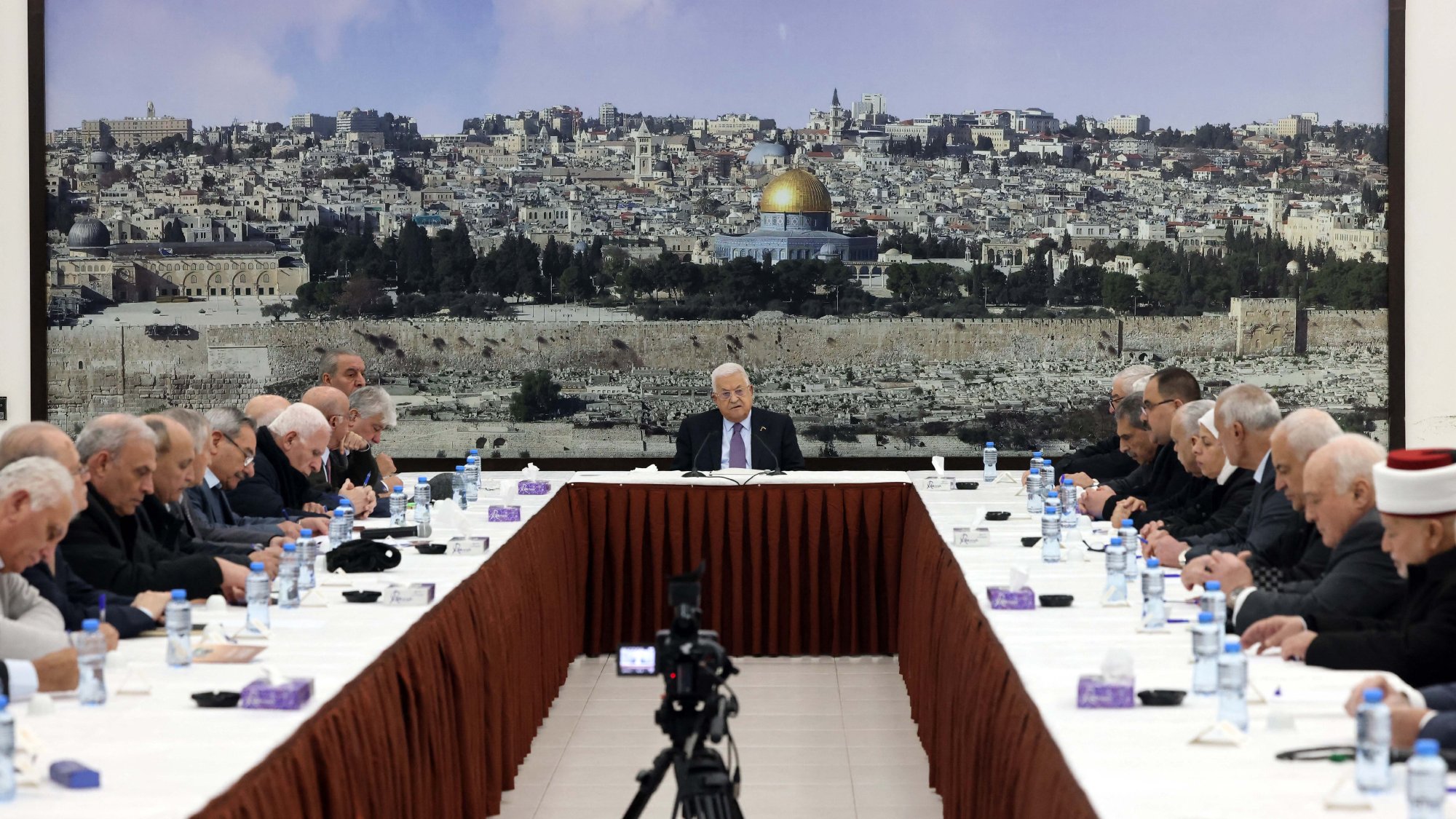 L'image montre une réunion se tenant autour d'une grande table ovale, avec plusieurs personnes assises de chaque côté. Au fond, il y a une grande image d'une ville, probablement Jérusalem, avec le dôme doré d'une mosquée bien visible. Les participants, qui semblent être des hommes âgés, portent des costumes sombres et affichent une expression sérieuse. Au centre de la table, une personne (probablement un chef ou un président) s'exprime pendant que les autres l'écoutent attentivement. Des bouteilles d'eau et des serviettes sont disposées sur la table. L'ambiance semble solennelle et attentive.