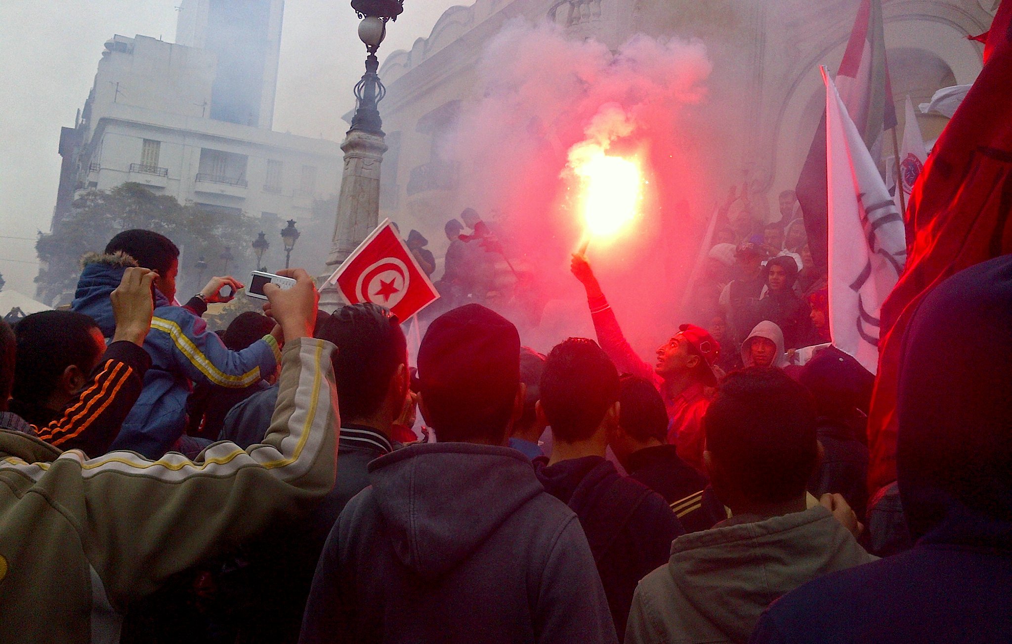 L'image montre une foule rassemblée dans une manifestation. Des personnes tiennent des drapeaux, dont un drapeau tunisien, et certains agitent des fumigènes qui dégagent de la fumée rouge. L'ambiance semble énergique et engagée, avec des bâtiments en arrière-plan. La foule paraît déterminée et unie autour de leur cause.