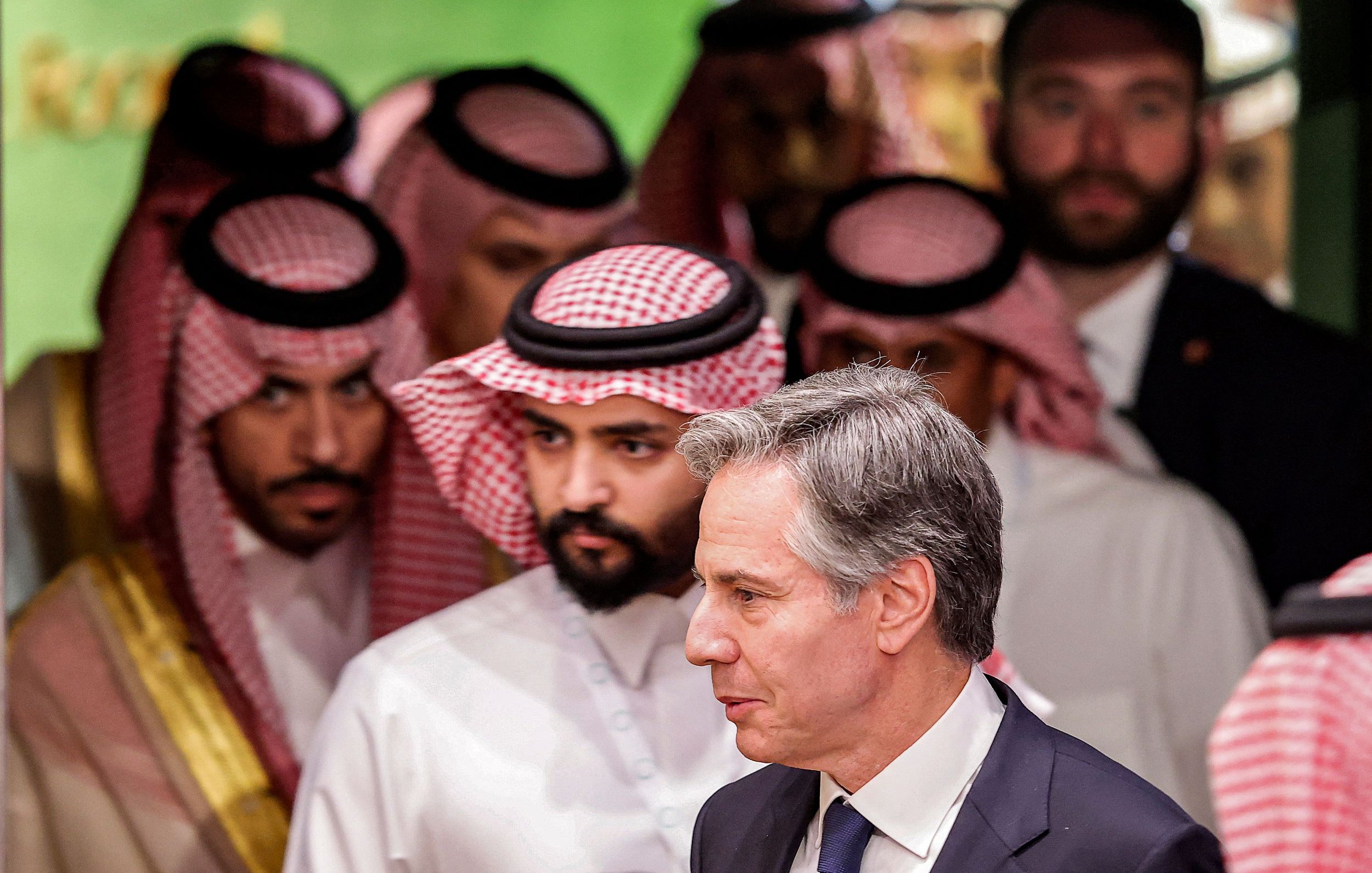 L'image montre un groupe d'hommes en vêtements traditionnels saoudiens, portant des ghoutas et des agals, en arrière-plan. Au premier plan, un homme en costume sombre marche, semblant engagé dans une conversation ou une réflexion. L'ambiance générale de l'image suggère une réunion ou un événement diplomatique.