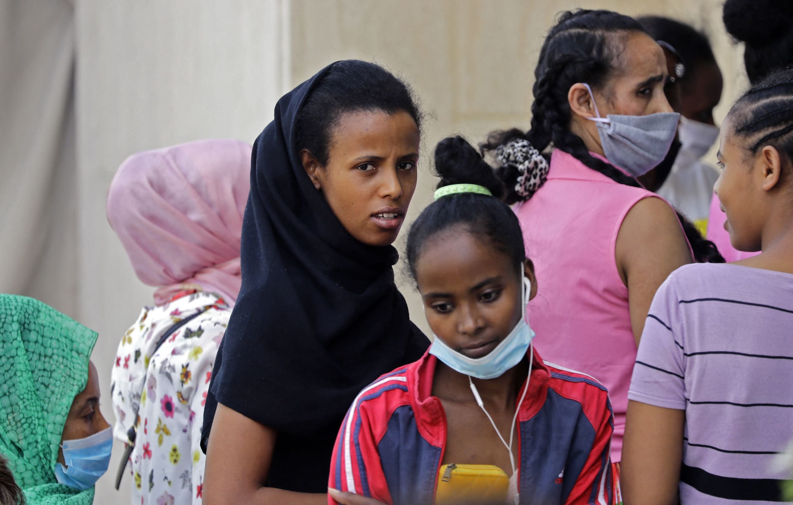 L'image montre un groupe de femmes et de jeunes filles, certaines portant des foulards ou des masques. Les visages expriment divers états d'esprit, allant de la concentration à l'inquiétude. Les vêtements sont colorés, et certaines d'entre elles semblent discuter ou interagir, tandis que d'autres regardent vers l'objectif avec curiosité. L'ambiance générale dénote un moment de rassemblement ou d'attente.