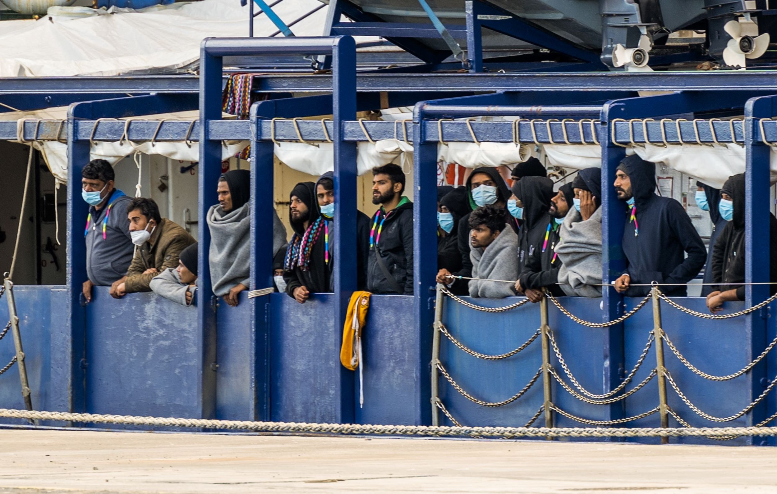 L'image montre un groupe de personnes se tenant sur un bateau. Elles portent des masques et des vêtements semblant chauds, comme des sweats à capuche. Les gens regardent vers l'extérieur, probablement vers la terre ou vers un port. L'environnement semble portuaire, avec des équipements et d'autres structures en arrière-plan. L'atmosphère peut sembler préoccupante ou attendue, reflétant une situation particulière.