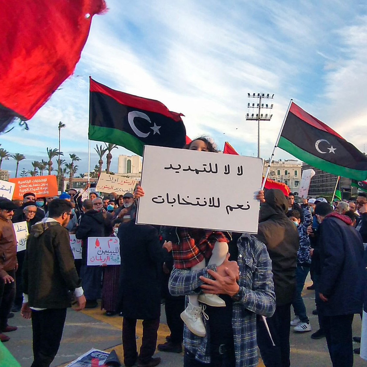 L'image montre une manifestation avec de nombreuses personnes rassemblées. Des drapeaux de la Libye flottent dans l'air, affichant des couleurs rouge, noir et vert. Au centre de la scène, une personne tient une pancarte avec des inscriptions en arabe. On peut également voir des participants portant des masques et d'autres pancartes, exprimant des revendications politiques. L'ambiance paraît dynamique, avec une multitude de personnes engagées dans une cause commune.