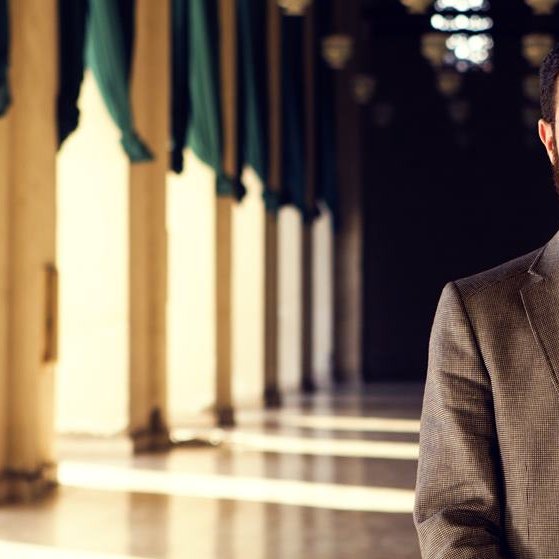 L'image montre un homme portant un costume avec une chemise à rayures. Il a une barbe bien entretenue et porte des lunettes. En arrière-plan, on peut voir un couloir avec des colonnes et des jeux d'ombres créés par la lumière qui entre. L'ambiance semble calme et chaleureuse, avec des tentures vertes accentuant l'architecture.