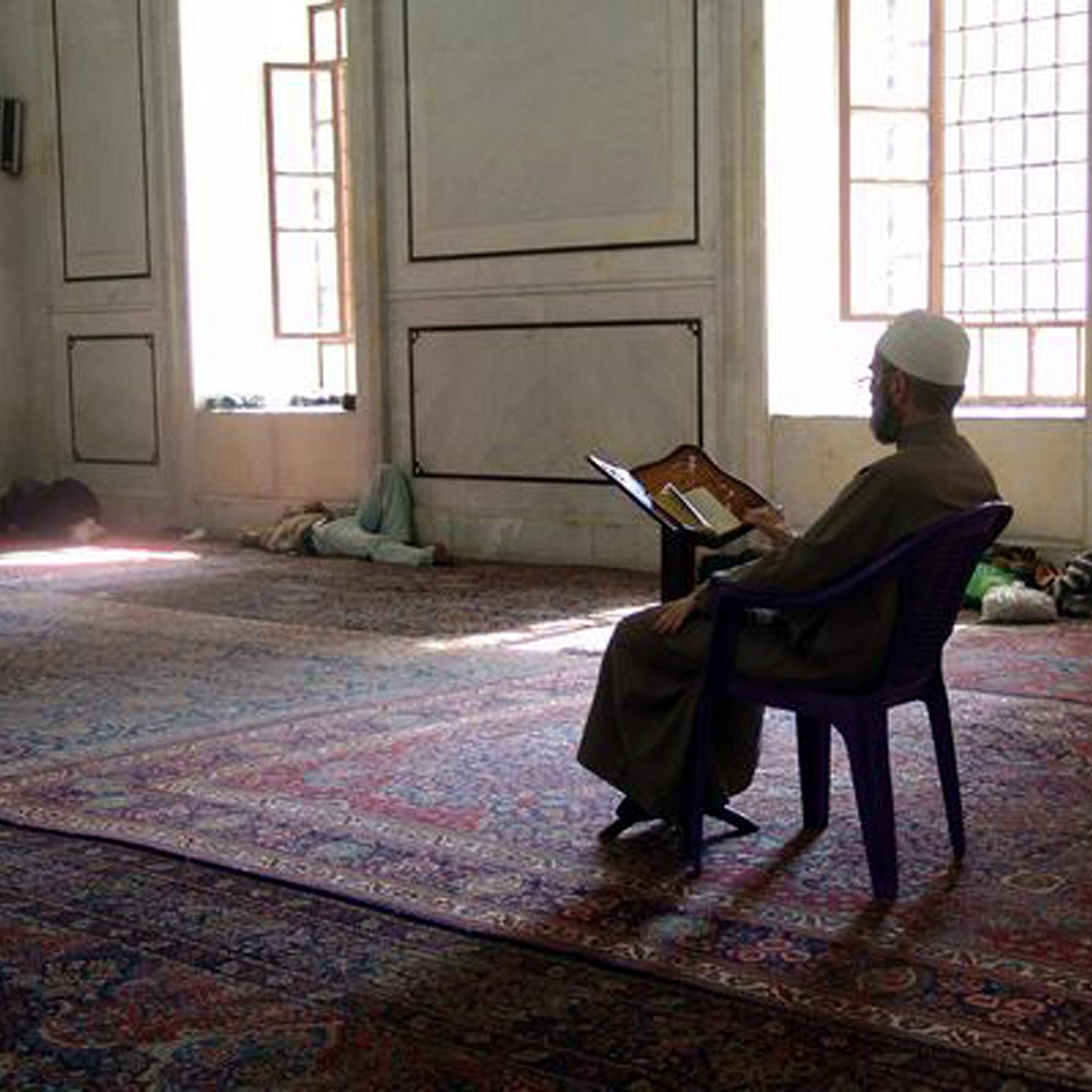 L'image montre un homme assis sur une chaise dans une mosquée. Il est vêtu d'une longue robe et porte un chapeau traditionnel. Devant lui, il lit un livre ouvert, probablement un exemplaire du Coran. La mosquée a un intérieur simple, avec des murs en marbre et des fenêtres laissant passer la lumière naturelle. Le sol est recouvert de tapis, et on peut voir d'autres personnes allongées au fond de la salle.