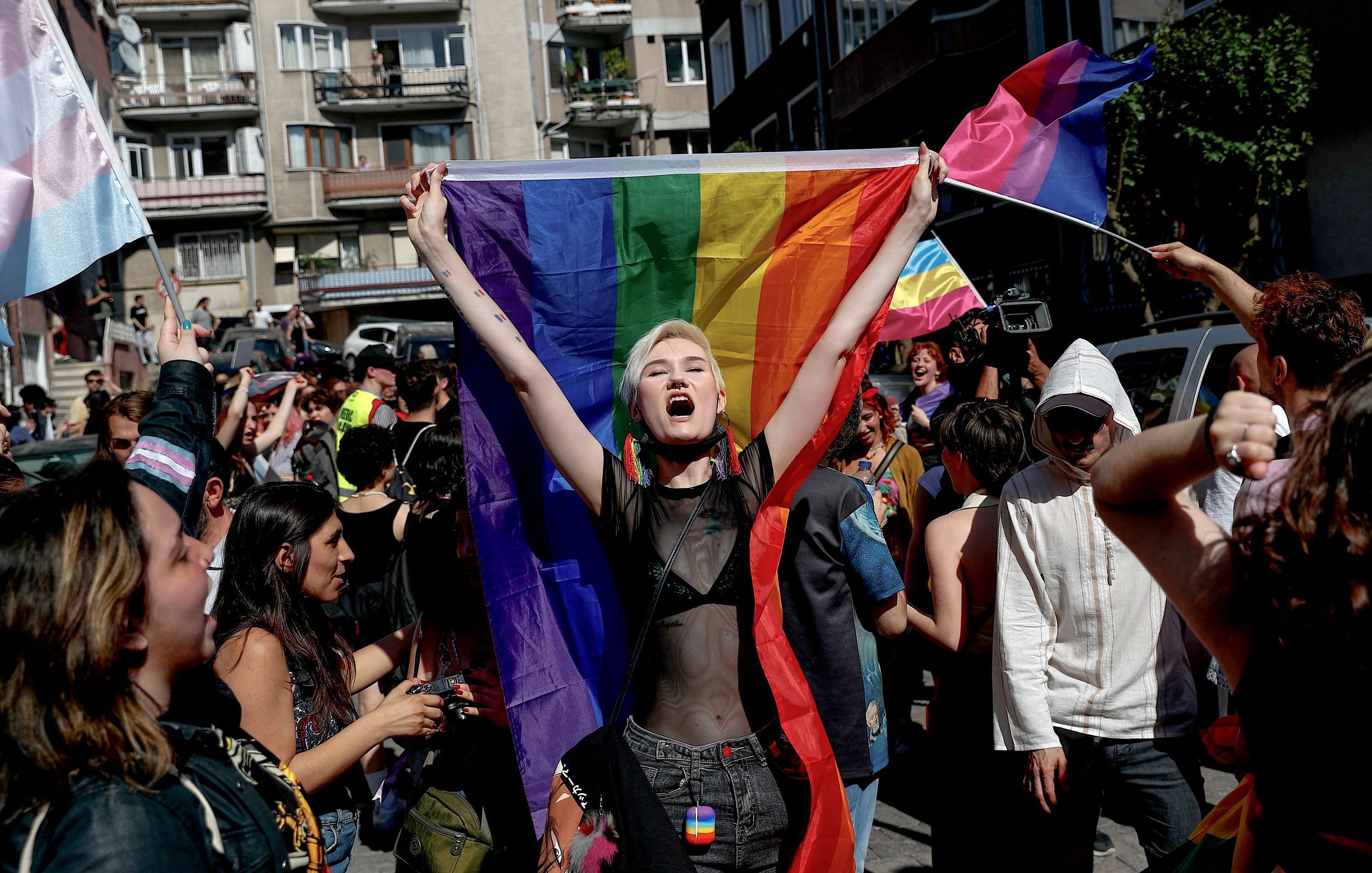 L'image montre une scène dynamique d'une marche ou d'une manifestation célébrant la fierté. Au centre, une personne tient une grande drapeau arc-en-ciel, symbole de la communauté LGBTQ+. Elle exprime sa joie ou son enthousiasme, visiblement impliquée dans l'événement. Autour d'elle, d'autres participants portent également des drapeaux et affichent des expressions de soutien et de célébration. L'environnement urbain en arrière-plan suggère que l'événement se déroule dans une ville. Les gens semblent heureux et engagés dans une atmosphère festive et inclusive.