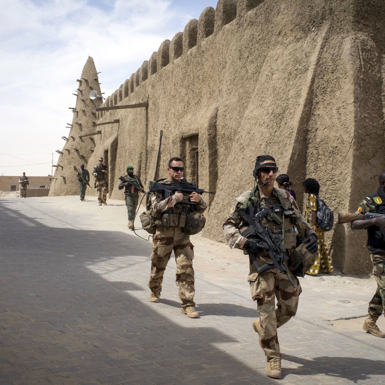 L'image montre une scène militaire dans une zone urbaine, probablement dans un pays du Sahel. Plusieurs soldats sont visibles, certains en uniforme camouflé et d'autres en tenue différente. Ils portent des armes et avancent dans une rue étroite bordée de bâtiments en terre battue, typiques de l'architecture sahélienne. On peut également apercevoir des civils en arrière-plan, ajoutant une dimension humaine à la scène. Le ciel est partiellement nuageux, ce qui suggère une ambiance chaude et aride.