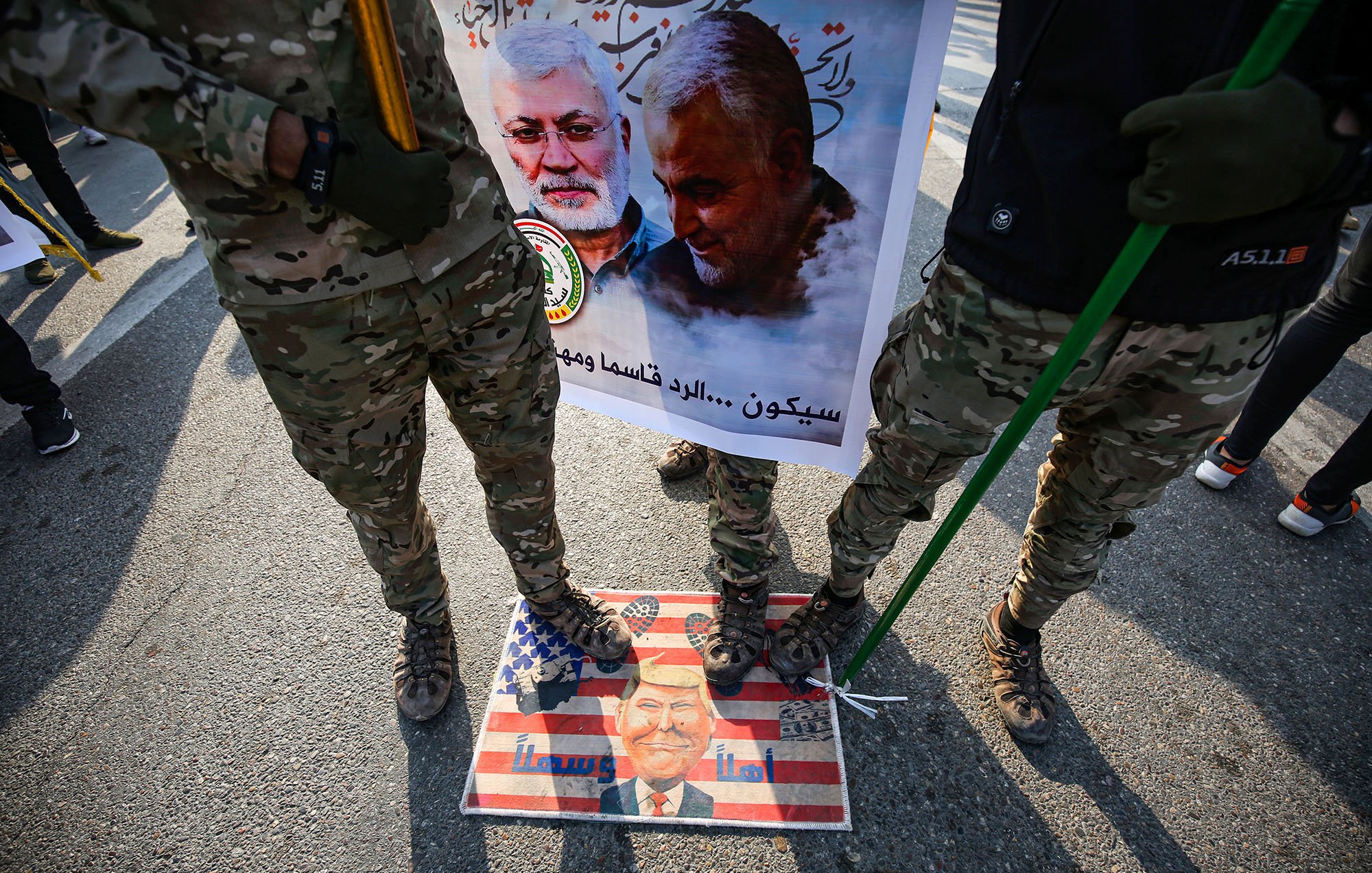 در این تصویر، تعدادی از افراد با لباس‌های نظامی و camo (رنگ‌های استتاری) در حال ایستادن هستند. آنها در دست خود عصا یا چوب‌هایی دارند. زیر پایشان تصویر یک پرچم آمریکاست که بر روی آن چهره‌ای در پایین مشاهده می‌شود. در پس‌زمینه، یک بنر بزرگ وجود دارد که شامل تصاویری از دو نفر است و متنی در مورد آنها نوشته شده است. فضایی حاکی از تظاهرات یا یک رویداد اجتماعی در حال شکل‌گیری است.