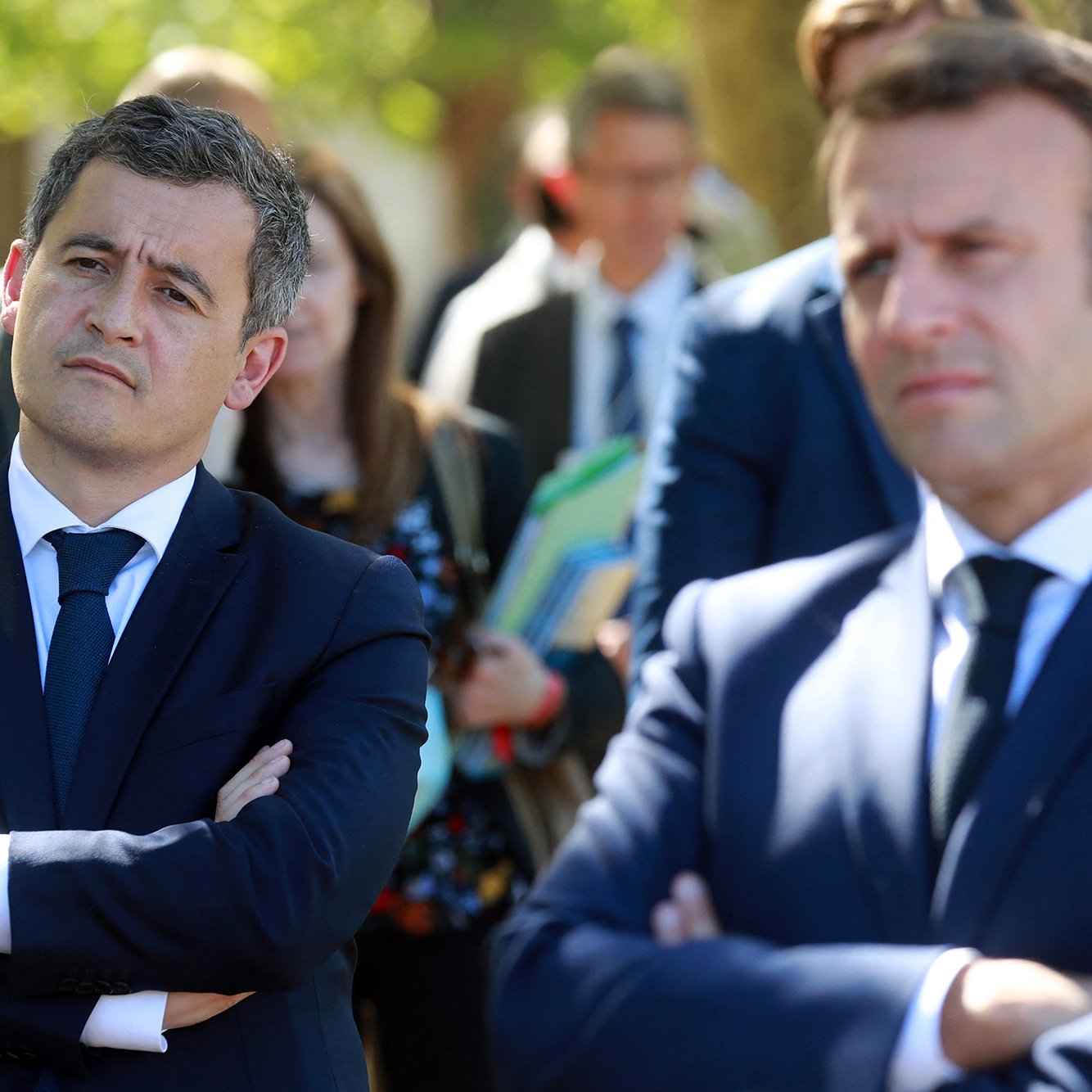 L'image montre deux hommes en costume, l'un avec les bras croisés et l'air pensif, et l'autre légèrement en avant, observant. Ils semblent participent à un événement public, entourés d'autres personnes en arrière-plan, dans un cadre extérieur. Les expressions faciales suggèrent qu'ils écoutent attentivement ou réfléchissent à ce qui se passe.