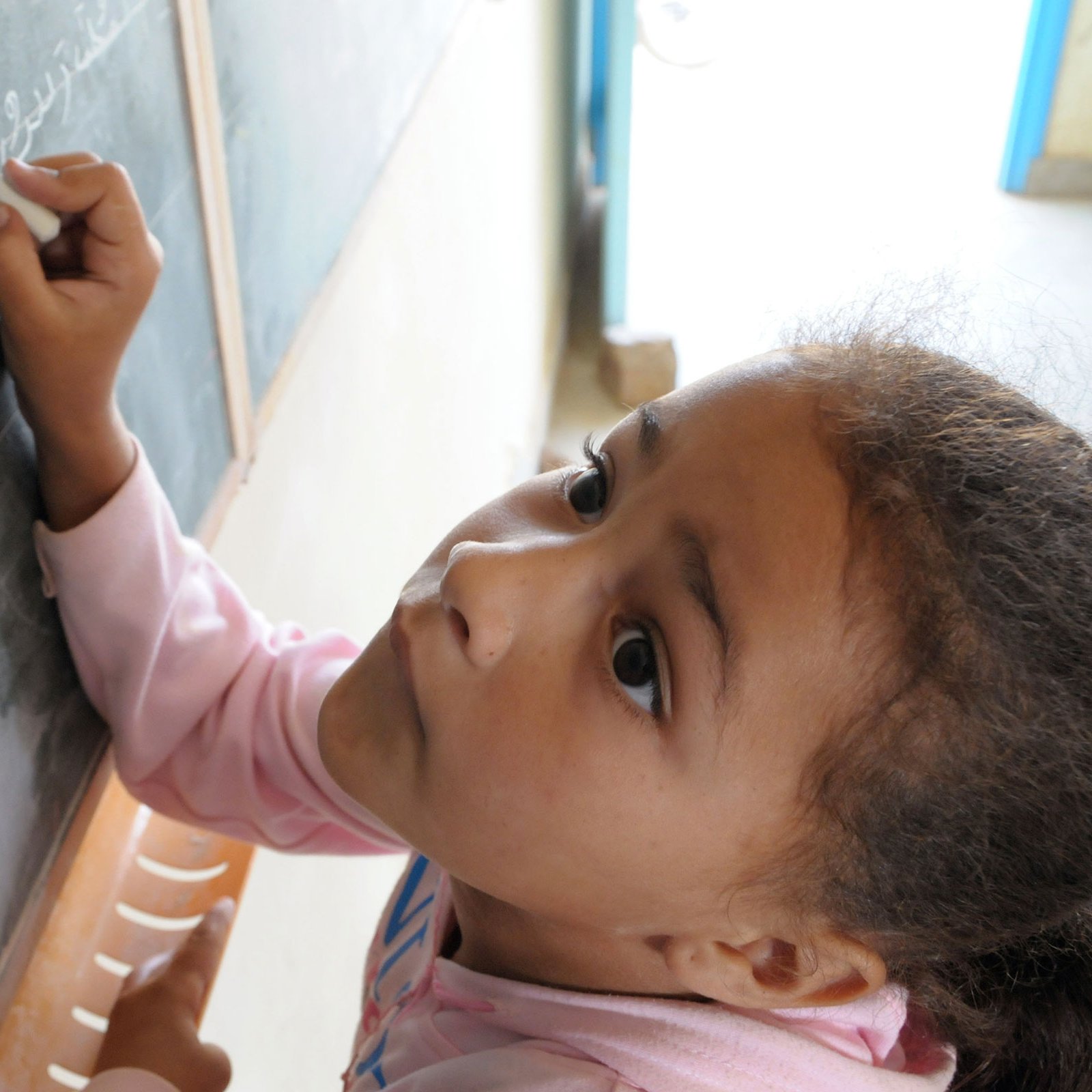 L'image montre une jeune fille penchée vers un tableau noir. Elle semble concentrée alors qu'elle écrit ou dessine quelque chose avec une craie. On peut voir des lettres ou des symboles tracés sur le tableau. La lumière entre par une fenêtre visible en arrière-plan, créant une ambiance chaleureuse. La fille porte un sweat à capuche rose et a les cheveux bouclés.