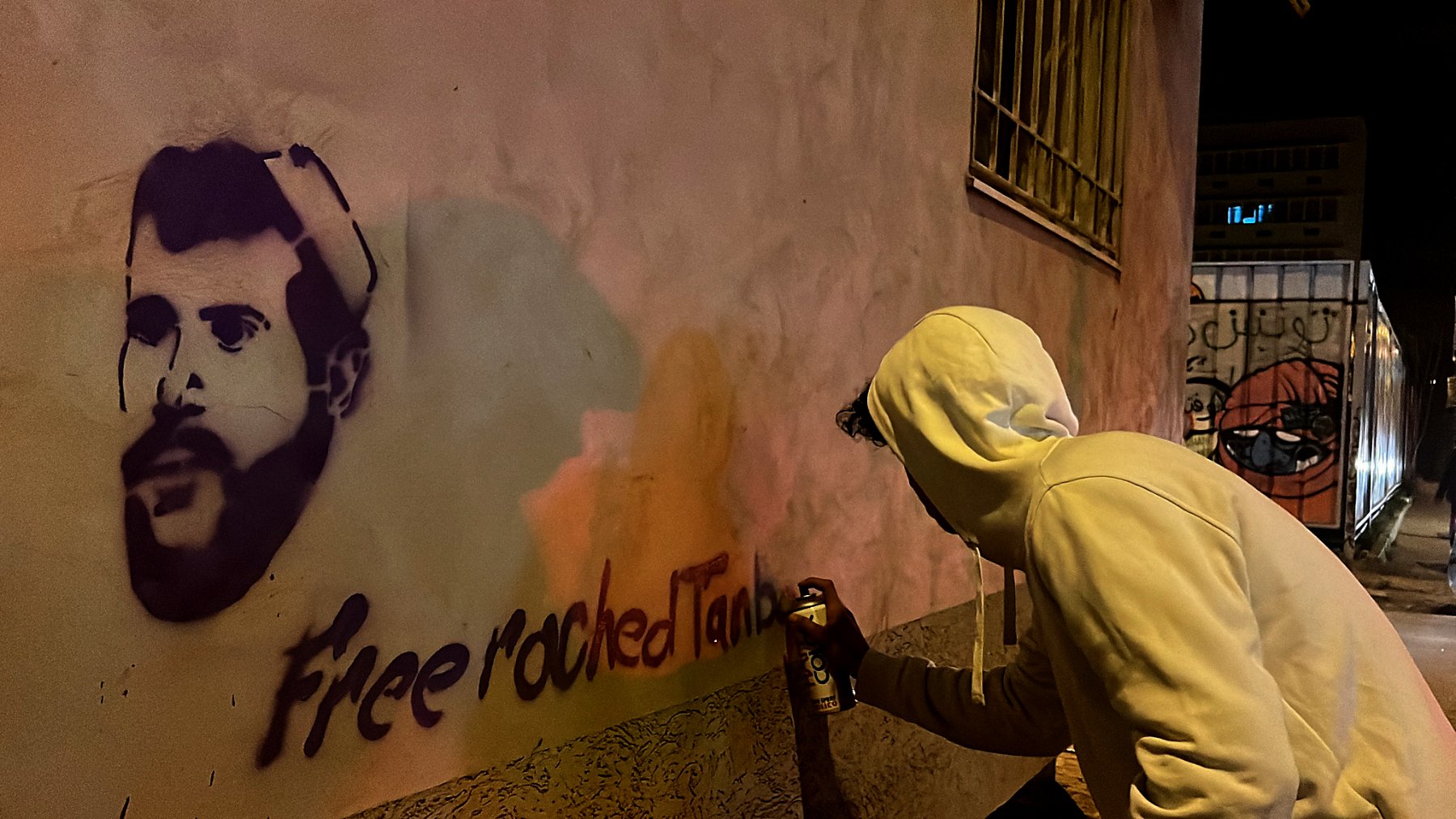 L'image montre une personne de dos, habillée d'un sweat à capuche blanc, en train de peindre un graffiti sur un mur. Le graffiti représente le visage d'un homme, probablement une figure connue, avec la phrase "Free roched Tank" écrite en dessous. La scène se déroule la nuit, et l'éclairage crée des reflets colorés sur le mur. On peut également apercevoir un portail ou une autre surface à proximité avec un autre graffiti en arrière-plan.