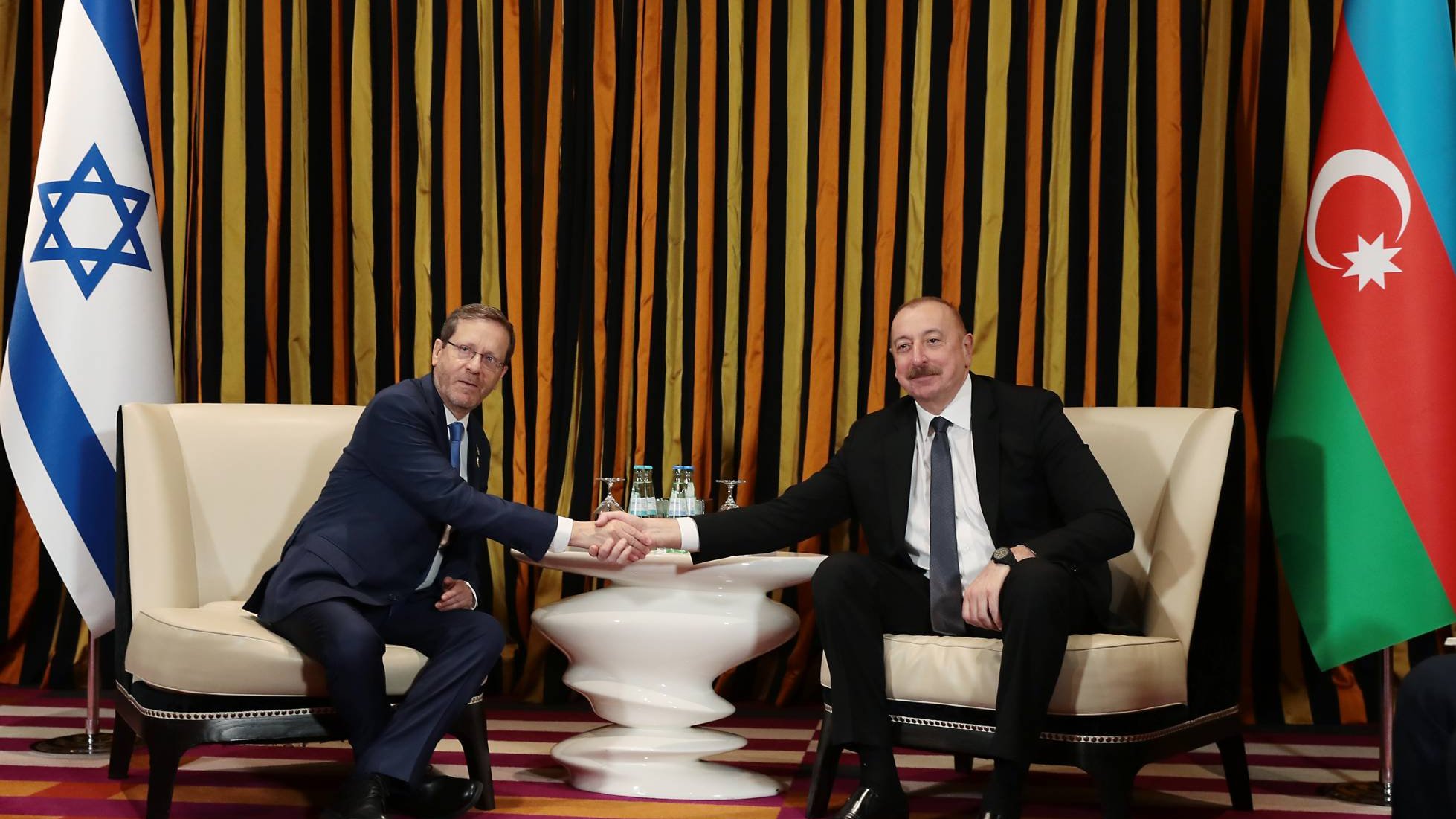 Deux hommes, en costume, se serrent la main dans un cadre élégant avec des drapeaux.