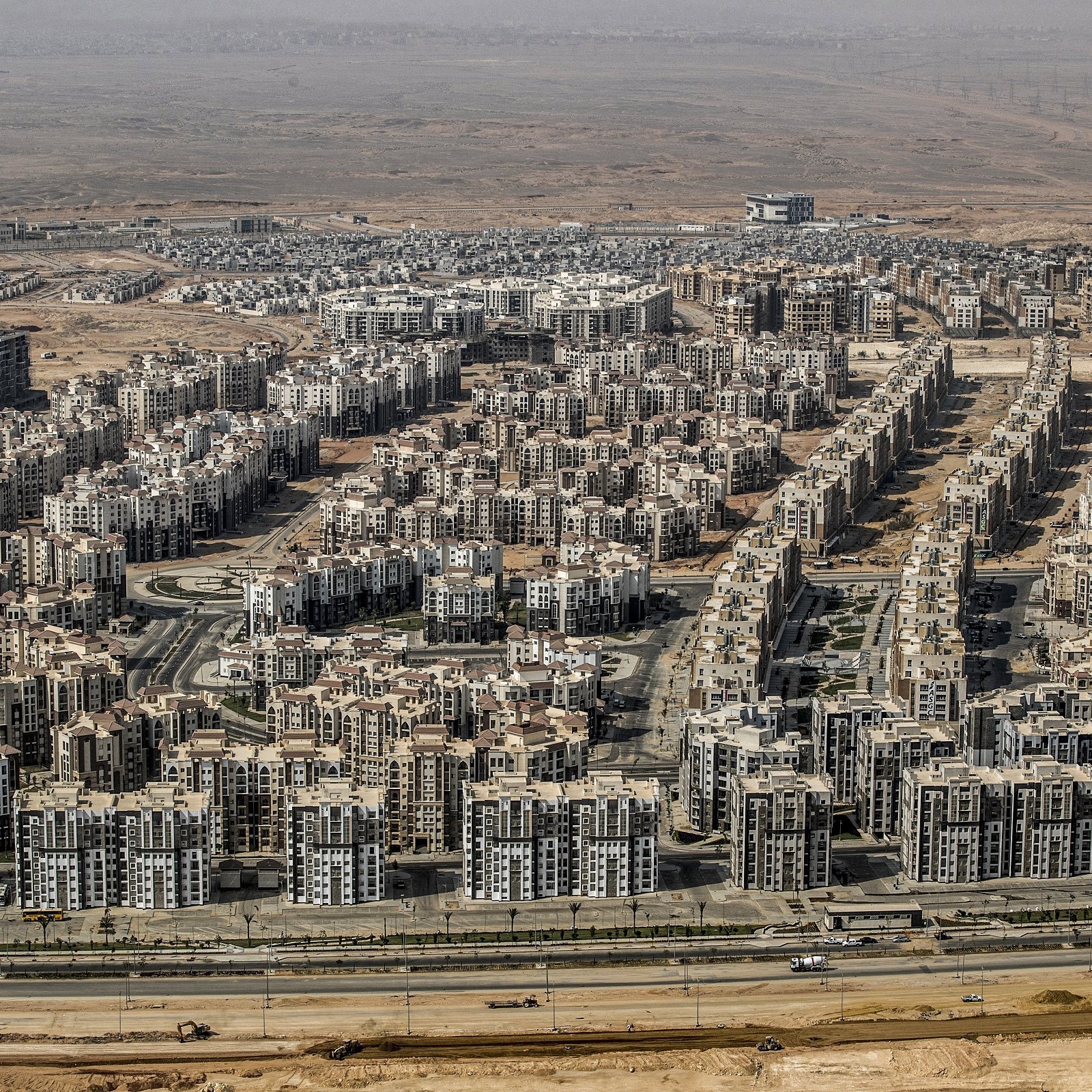 L'image montre une vaste zone urbaine composée de nombreux immeubles de plusieurs étages, organisés de manière dense. On peut voir des routes qui traversent ces secteurs résidentiels, avec des infrastructures modernes. En arrière-plan, le paysage est désertique, ce qui évoque un contraste entre l'urbanisation et l'environnement naturel environnant. L'ensemble dégage une impression de développement urbain important dans une région aride.