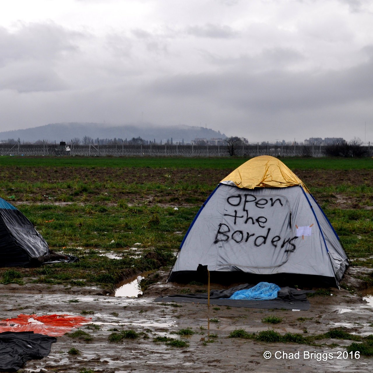 L'image montre un campement de tentes installé dans un terrain boueux. On aperçoit plusieurs tentes, dont l'une a un message écrit : "Open the Borders". Le paysage environnant est composé de champs et d'une arrière-plan nuageux, ce qui crée une atmosphère morose. Le sol est détrempé, indiquant probablement des conditions météorologiques difficiles.
