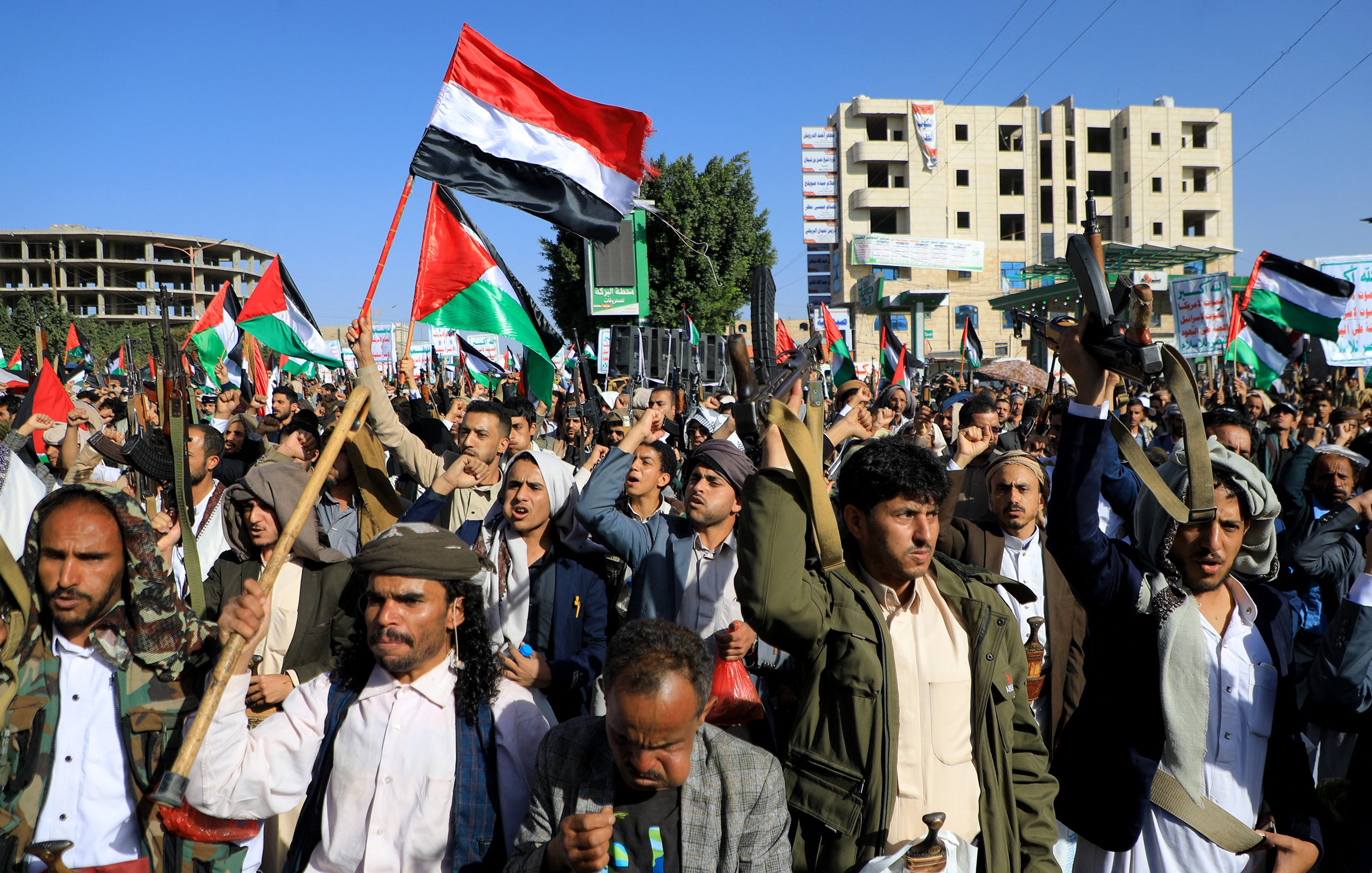 L'immagine mostra una folla di persone in protesta, che si riunisce in una manifestazione. I manifestanti alzano bandiere, probabilmente di carattere nazionale o politico, e sembrano esprimere il loro fervore con entusiasmo. Molti indossano abiti tradizionali e ci sono anche elementi di armi, come fucili, che suggeriscono una manifestazione di forte impatto emotivo e potenzialmente conflittuale. Gli edifici sullo sfondo indicano un contesto urbano, mentre l'atmosfera complessiva sembra intensa e carica di significato.