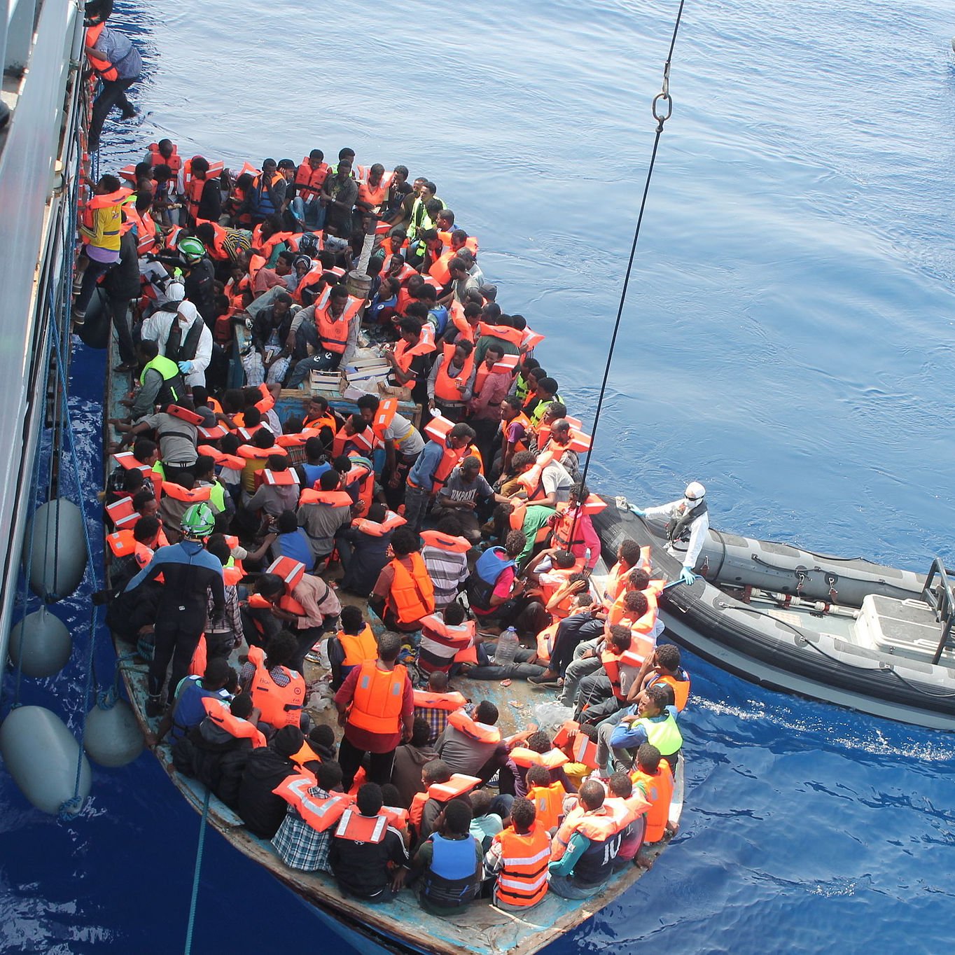 L'image montre un groupe de personnes dans une embarcation sur une mer calme. La plupart des individus portent des gilets de sauvetage orange, indiquant qu'ils sont en situation de danger. À proximité, une autre petite embarcation semble aider le groupe, probablement une équipe de secours. L'atmosphère générale évoque des enjeux humanitaires, liés à la migration et aux sauvetages en mer. Le ciel est clair et l'eau est d'un bleu profond.