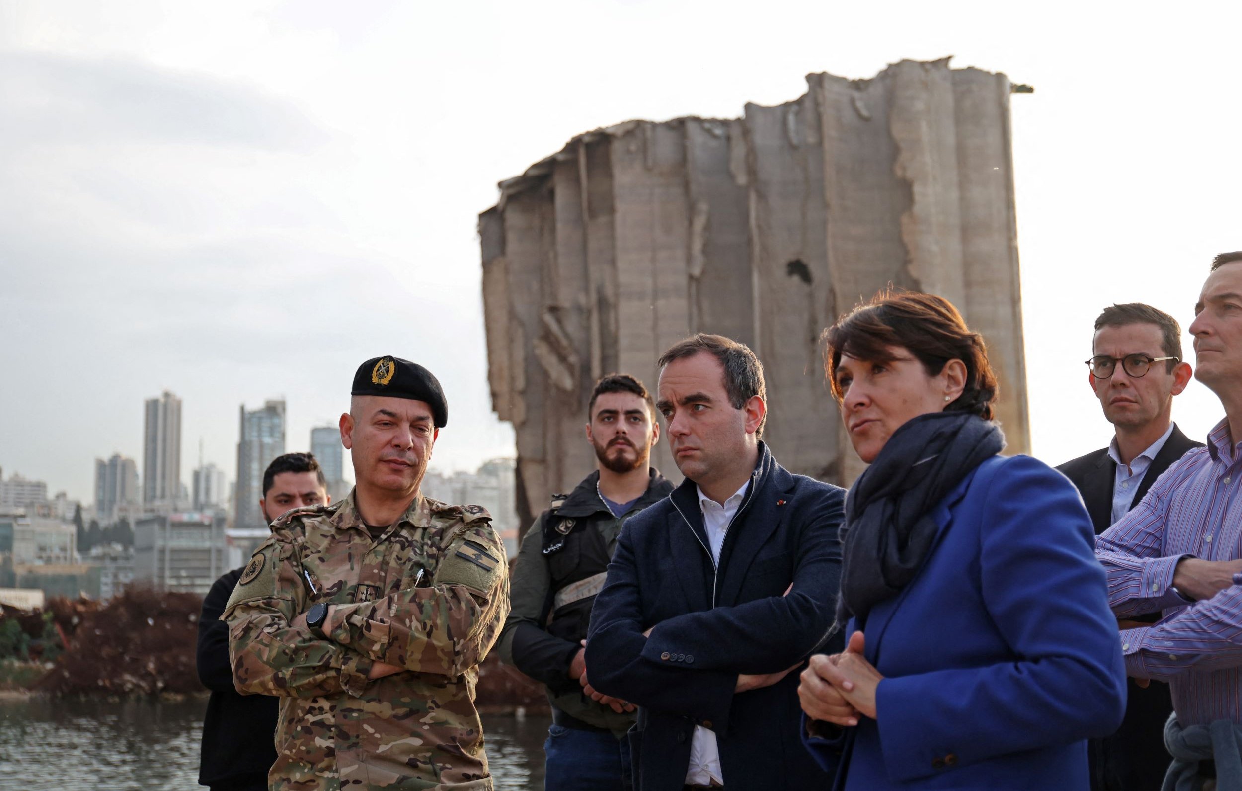 L'image montre plusieurs personnes rassemblées devant une structure endommagée, probablement un bâtiment ou un silo, avec un paysage urbain en arrière-plan. Parmi eux, on distingue un militaire en uniforme, entouré d'autres individus, dont des hommes et des femmes, portant des vêtements civils. L'ambiance semble sérieuse, comme si une discussion ou une évaluation de la situation était en cours. La lumière du jour diminue, suggérant que c'est en fin d'après-midi ou en début de soirée.