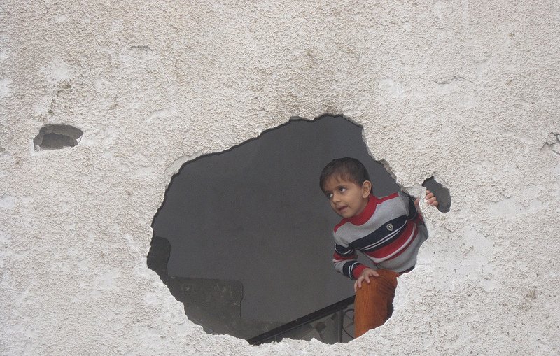 L'image montre un enfant qui regarde à travers un trou dans un mur. Le mur est en mauvais état, avec une surface rugueuse et des fissures visibles. L'enfant porte un pull rayé et semble curieux, se penchant légèrement pour observer ce qui se trouve de l'autre côté du mur. La scène évoque une atmosphère d'exploration et d'intrigue.
