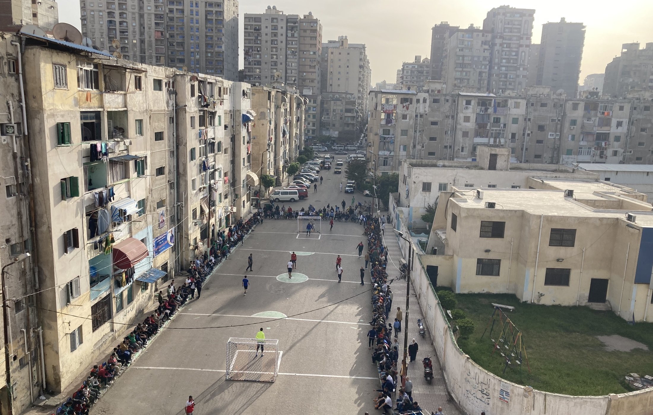 L'image montre un terrain de football encadré par des immeubles d'apartements dans une zone urbaine. Plusieurs personnes sont rassemblées sur des bancs le long du terrain, probablement en train de regarder un match. On peut voir des joueurs en action sur le terrain. L'établissement est entouré de bâtiments de plusieurs étages, témoignant d'une ambiance animée et communautaire. L'éclairage du soir ajoute une atmosphère chaleureuse à la scène.