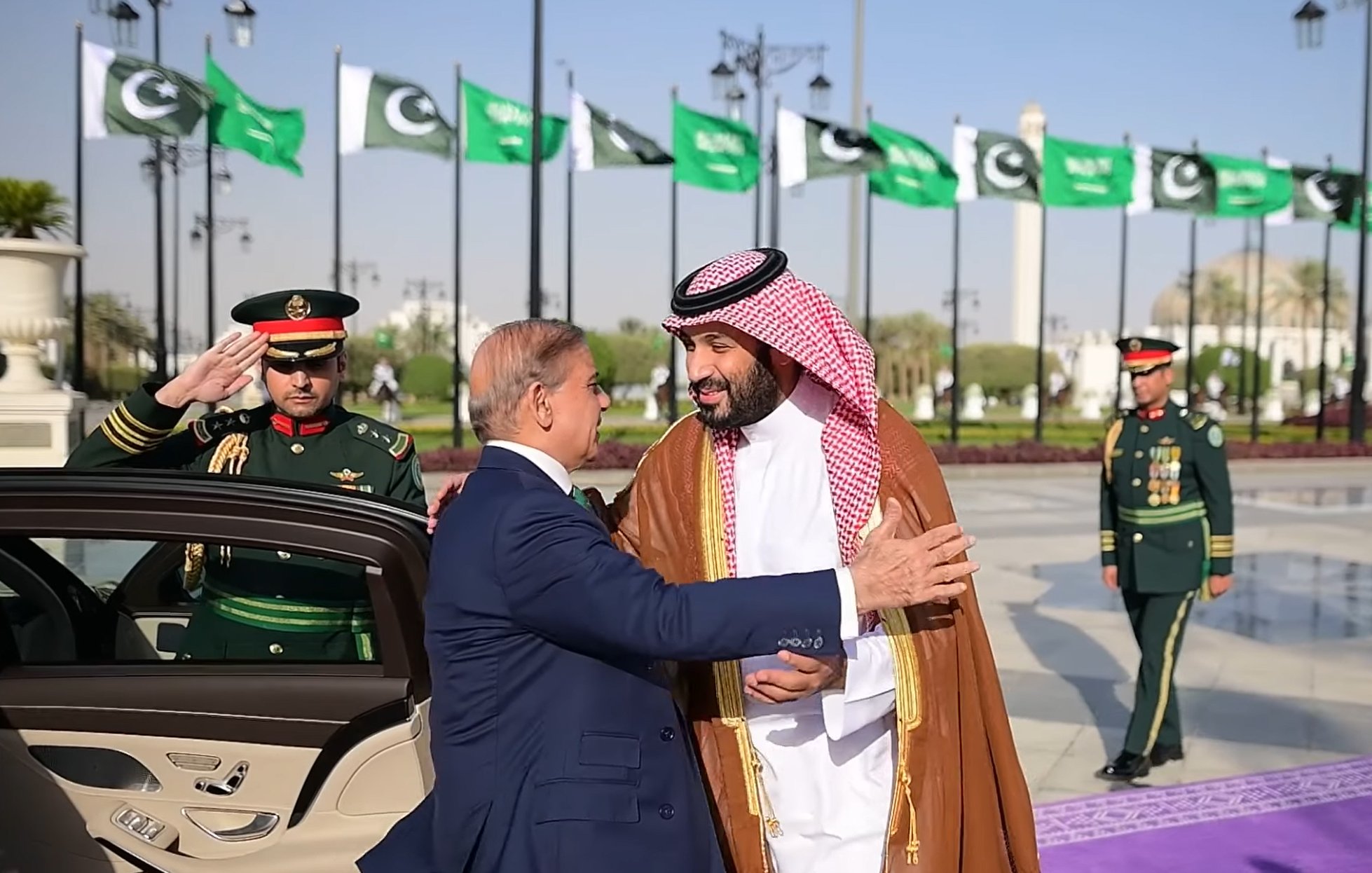 Deux hommes en costume échangent une accolade, avec des drapeaux en arrière-plan.