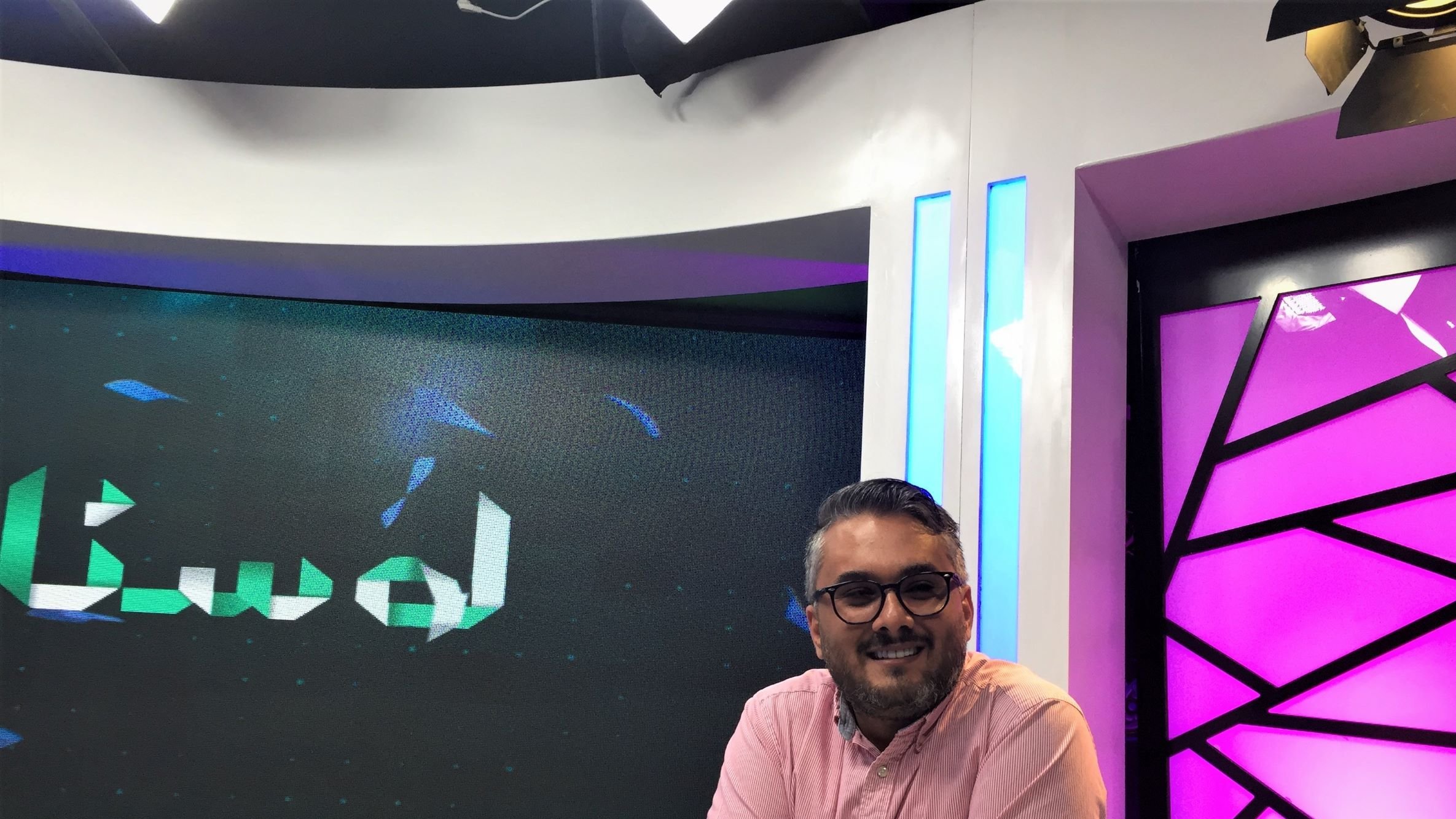 L'immagine mostra un uomo sorridente seduto davanti a una scrivania in uno studio televisivo. Indossa una camicia rosa e presenta un aspetto rilassato e accogliente. Dietro di lui si possono notare schermi che mostrano un logo o un testo in arabo, mentre l'illuminazione dello studio è accentuata da luci colorate, in particolare tonalità di viola e blu. L'ambiente sembra moderno e professionale, tipico di una trasmissione televisiva.