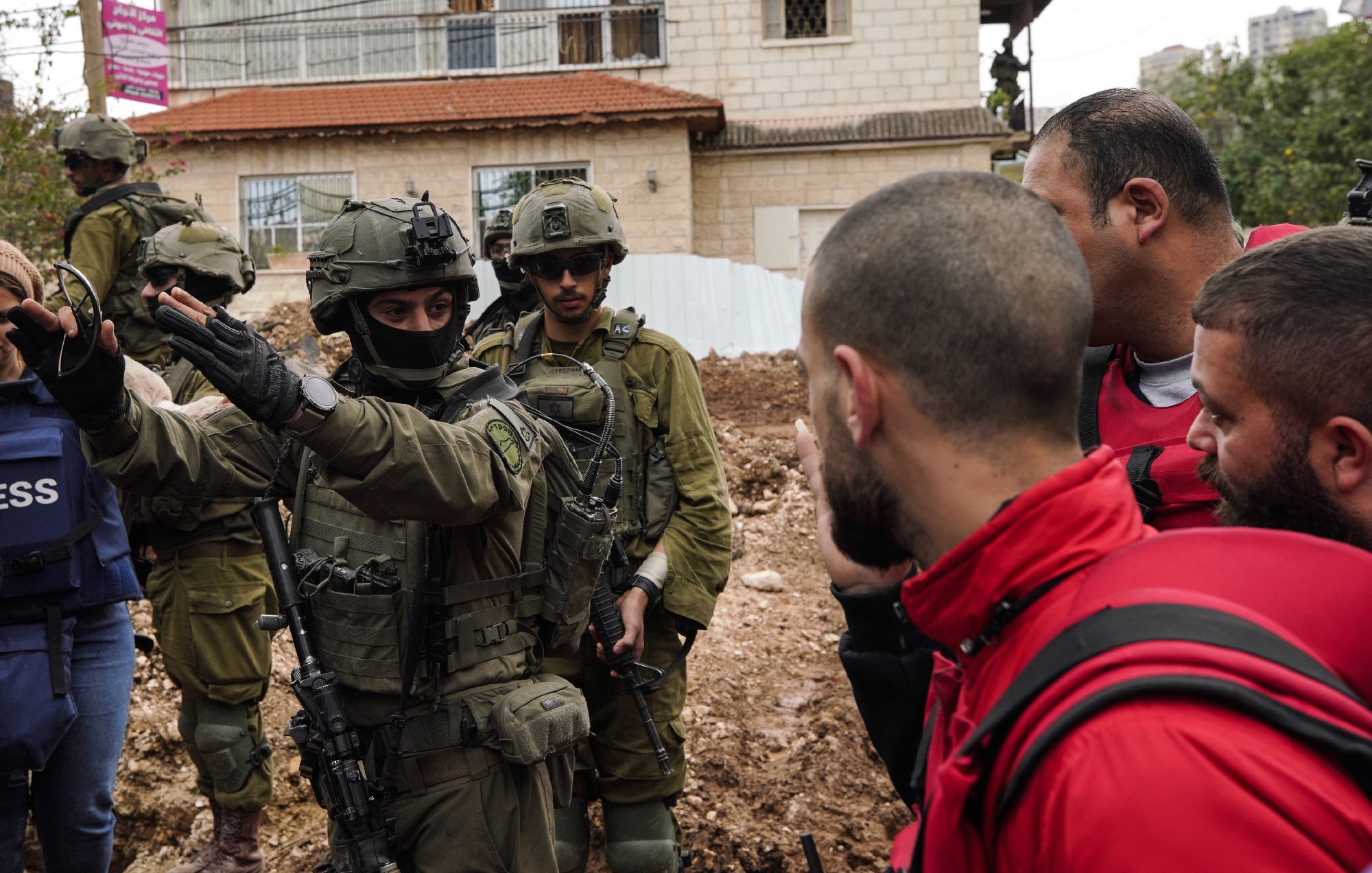 L'image montre des soldats en uniforme militaire, armés, entourés par un groupe de personnes. Certains sont en tenue d'urgence, tandis que d'autres semblent discuter avec les soldats. En arrière-plan, on voit des bâtiments et un terrain boueux. L'ambiance est tendue.