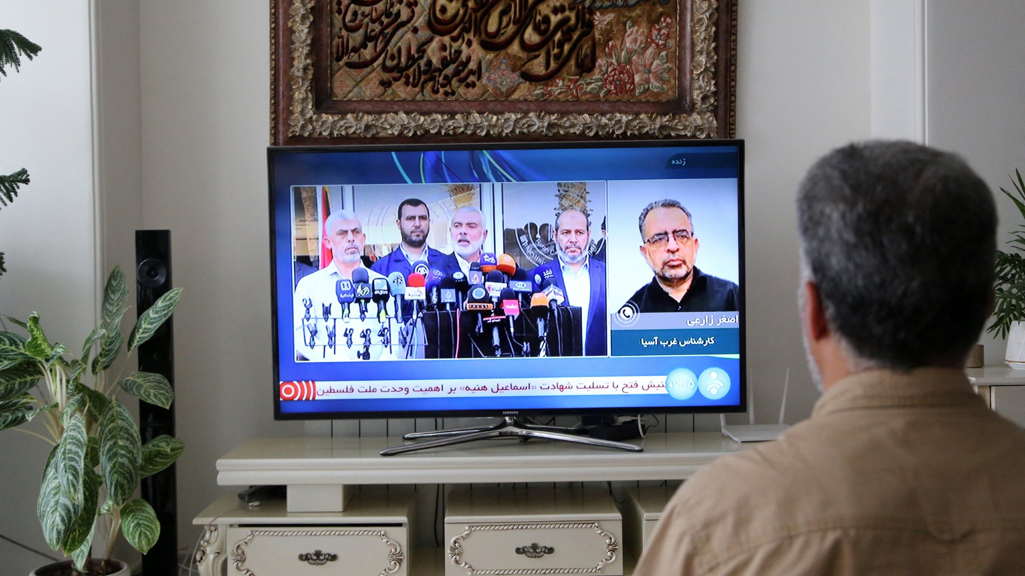A person is sitting on a couch, facing a television that is displaying a news broadcast featuring several speakers at a press conference. In the background, there is a decorative wall hanging and a plant to the side.