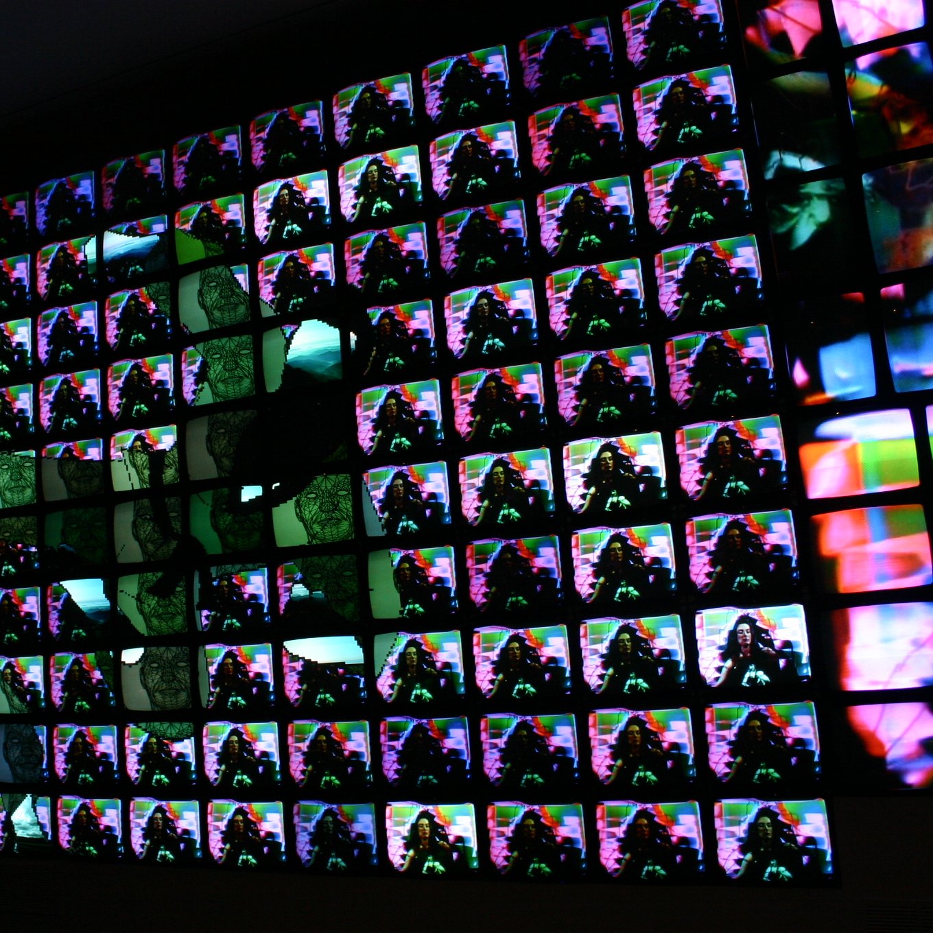 L'image présente une installation artistique composée de nombreux écrans alignés de manière à former un tableau visuel. Chaque écran affiche des images colorées, vibrantes et abstraites, créant un effet d'ensemble dynamique. Les couleurs se mélangent et se chevauchent, ajoutant une sensation de mouvement. La composition semble jouer avec la lumière et les contrastes, attirant l'œil vers des motifs diversifiés. L'ensemble évoque une immersion dans une expérience numérique moderne et artistique.