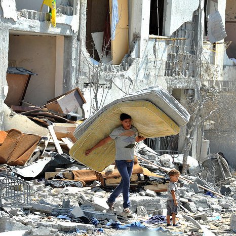 L'image montre un jeune homme portant un matelas sur son dos, marchant à travers des décombres. À côté de lui, un petit garçon l'observe. Le décor est celui d'un bâtiment en ruines, avec des meubles éparpillés et des murs endommagés, ce qui suggère une scène de destruction et de détresse. L'atmosphère est empreinte de tristesse et de désolation.