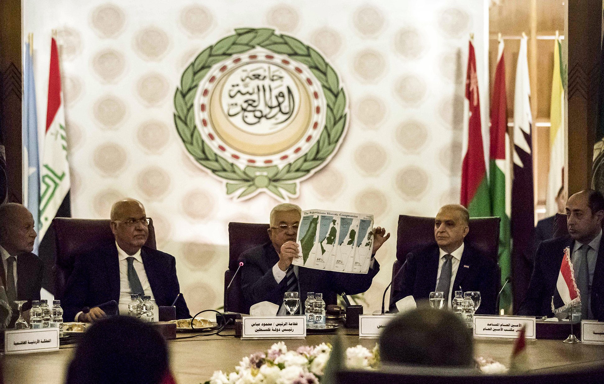 L'image montre une réunion officielle, probablement dans le cadre d'une organisation internationale. Plusieurs hommes sont assis autour d'une table dans un espace bien décoré, avec des drapeaux de différents pays en arrière-plan. L'un des hommes, au centre, tient un document ou un graphique qu'il montre aux autres participants. Les autres semblent attentifs et engagés dans la discussion. Les éléments visuels suggèrent une atmosphère sérieuse et formelle, en lien avec des discussions diplomatiques.
