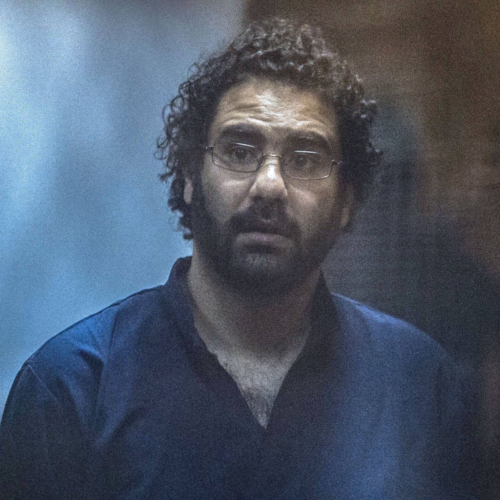 L'immagine mostra un uomo con capelli ricci e occhiali. Indossa una maglietta o camicia scura, ed appare sorpreso o preoccupato. Lo sfondo è sfocato e contribuisce a un'atmosfera di tensione o intensità. L'illuminazione è tenue, dando un effetto drammatico alla scena.