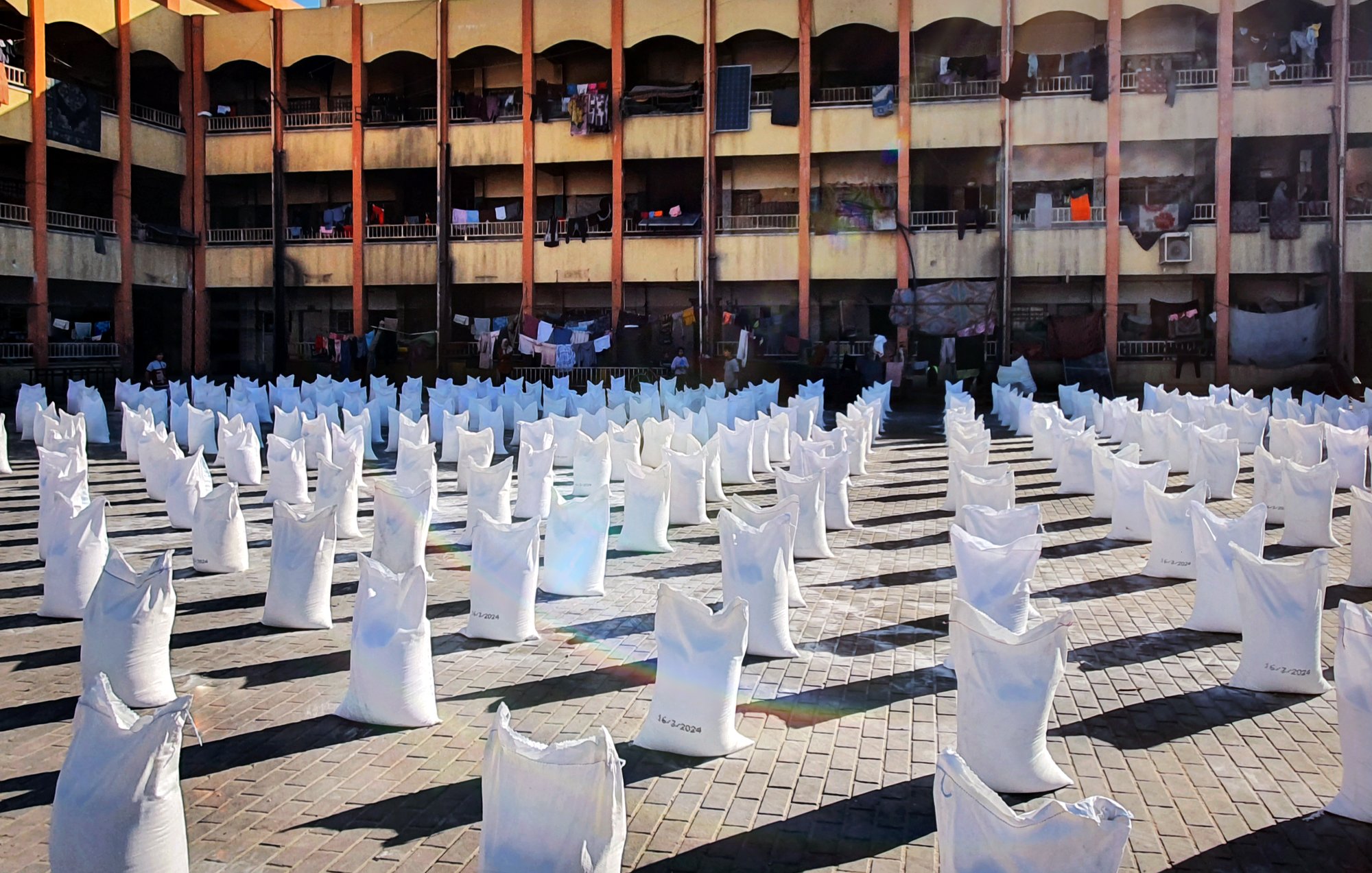 L'image représente un espace ouvert où de nombreux sacs blancs sont disposés en lignes. Ces sacs semblent être remplis de quelque chose, et le sol est constitué de pavés. En arrière-plan, on peut voir un bâtiment à plusieurs étages avec des balcons. Des vêtements sont suspendus à l'extérieur des balcons, suggérant une utilisation résidentielle de cet espace. La lumière du soleil éclaire la scène, créant un contraste entre les sacs blancs et l'environnement.