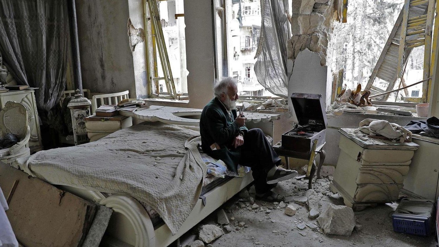 L'image montre un espace intérieur dévasté, probablement un appartement. Au centre, un homme âgé est assis sur un lit, tenant un objet à la main, peut-être une cigarette. L'environnement est marqué par des murs endommagés, des débris éparpillés et des fenêtres brisées, laissant voir l'extérieur. La lumière naturelle pénètre par les ouvertures, révélant l'état de désolation de la pièce. L'ambiance est sombre et mélancolique, illustrant les conséquences d'un conflit ou d'une catastrophe.