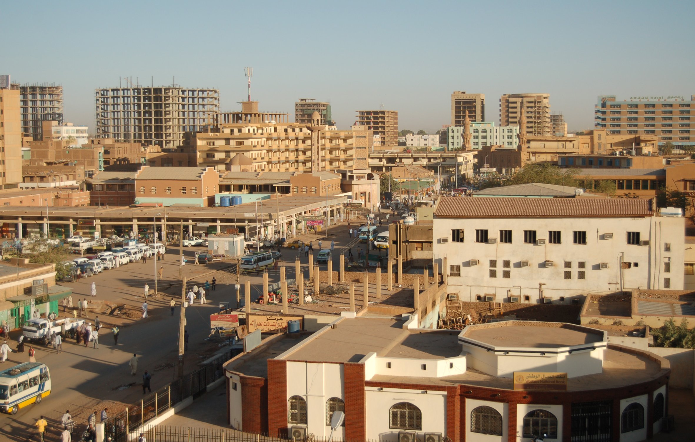 L'image montre une vue panoramique d'une ville, probablement une ville nord-africaine ou du Moyen-Orient. On peut voir plusieurs bâtiments, certains modernes et d'autres plus anciens, ainsi que des structures en construction. La rue est animée avec des voitures et des gens qui se déplacent. L'architecture varie, avec un mélange de styles. Le ciel est dégagé, indiquant probablement une belle journée ensoleillée. Les bâtiments sont de différentes hauteurs et couleurs, donnant à la scène un aspect dynamique et vivant.