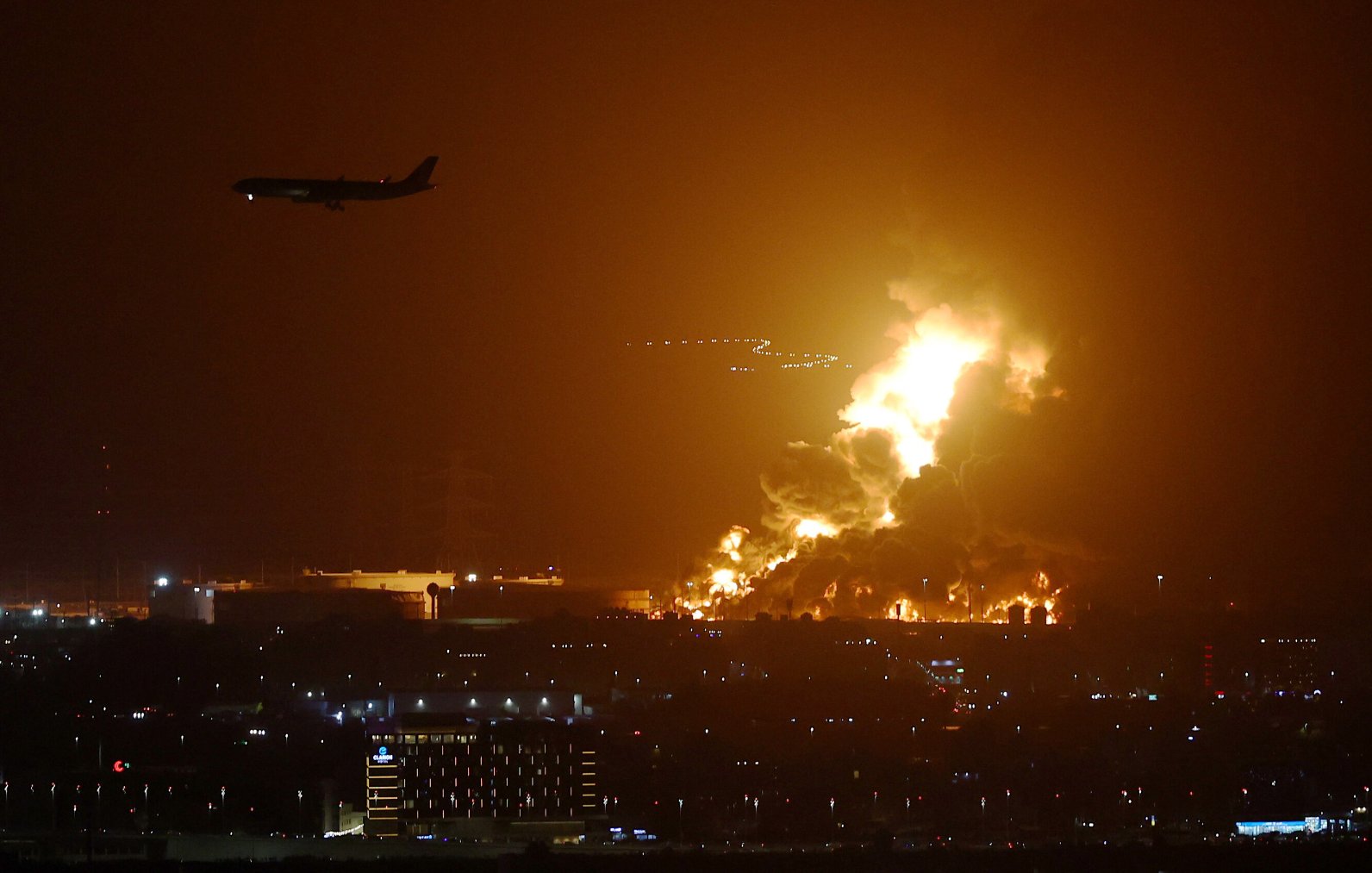 L'image montre un paysage nocturne éclairé par des flammes intenses, suggérant un incendie ou une explosion. On peut voir des colonnes de feu se levant dans le ciel, créant une ambiance dramatique. À l'arrière-plan, un avion est en train de survoler la scène, ce qui indique la proximité d'un aéroport. En bas, des lumières de la ville sont visibles, ajoutant un contraste entre l'urbanité et la catastrophe en cours.