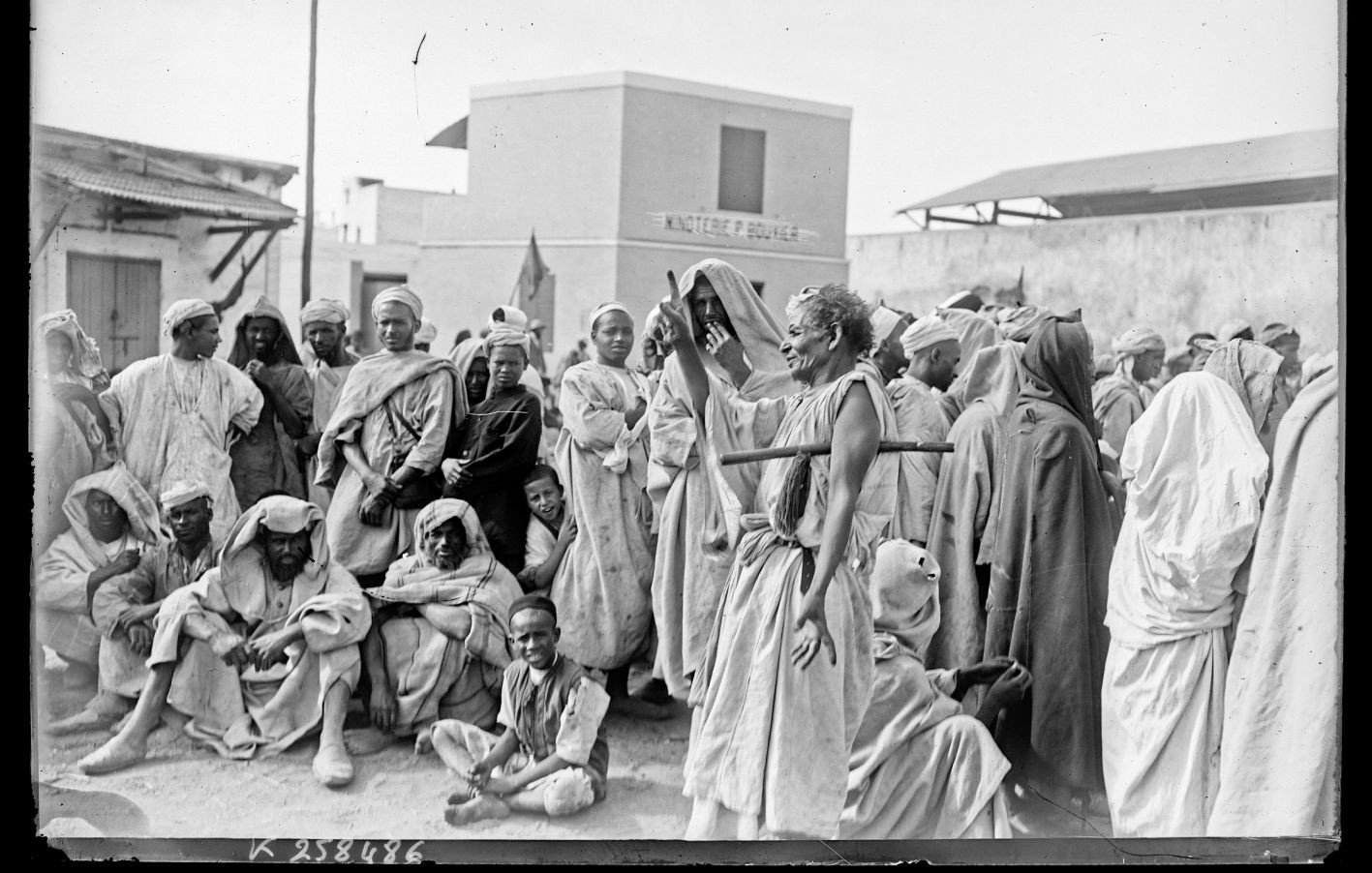 L'image montre un groupe de personnes rassemblées dans un environnement extérieur. Elles portent des vêtements traditionnels et semblent discuter ou observer quelque chose au centre. L'atmosphère est marquée par une certaine sérénité, mais aussi par une curiosité collective. En arrière-plan, on peut distinguer des bâtiments, suggérant un lieu structuré, potentiellement un marché ou un rassemblement communautaire. La lumière naturelle éclaire la scène, ajoutant à l'authenticité de l'ambiance.